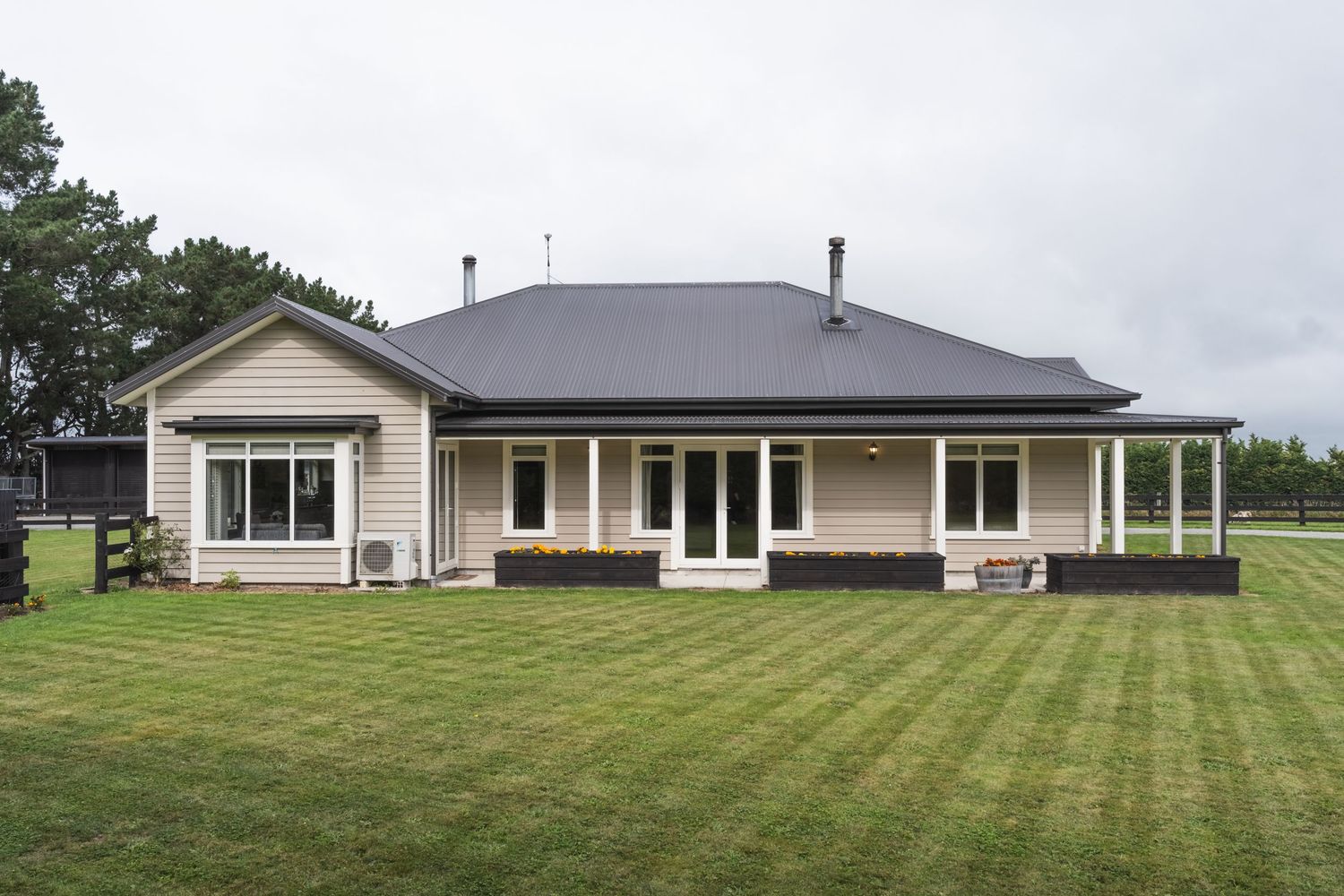Single-story beige farmhouse with dark roof, wraparound porch, and lush green lawn on a cloudy day.
