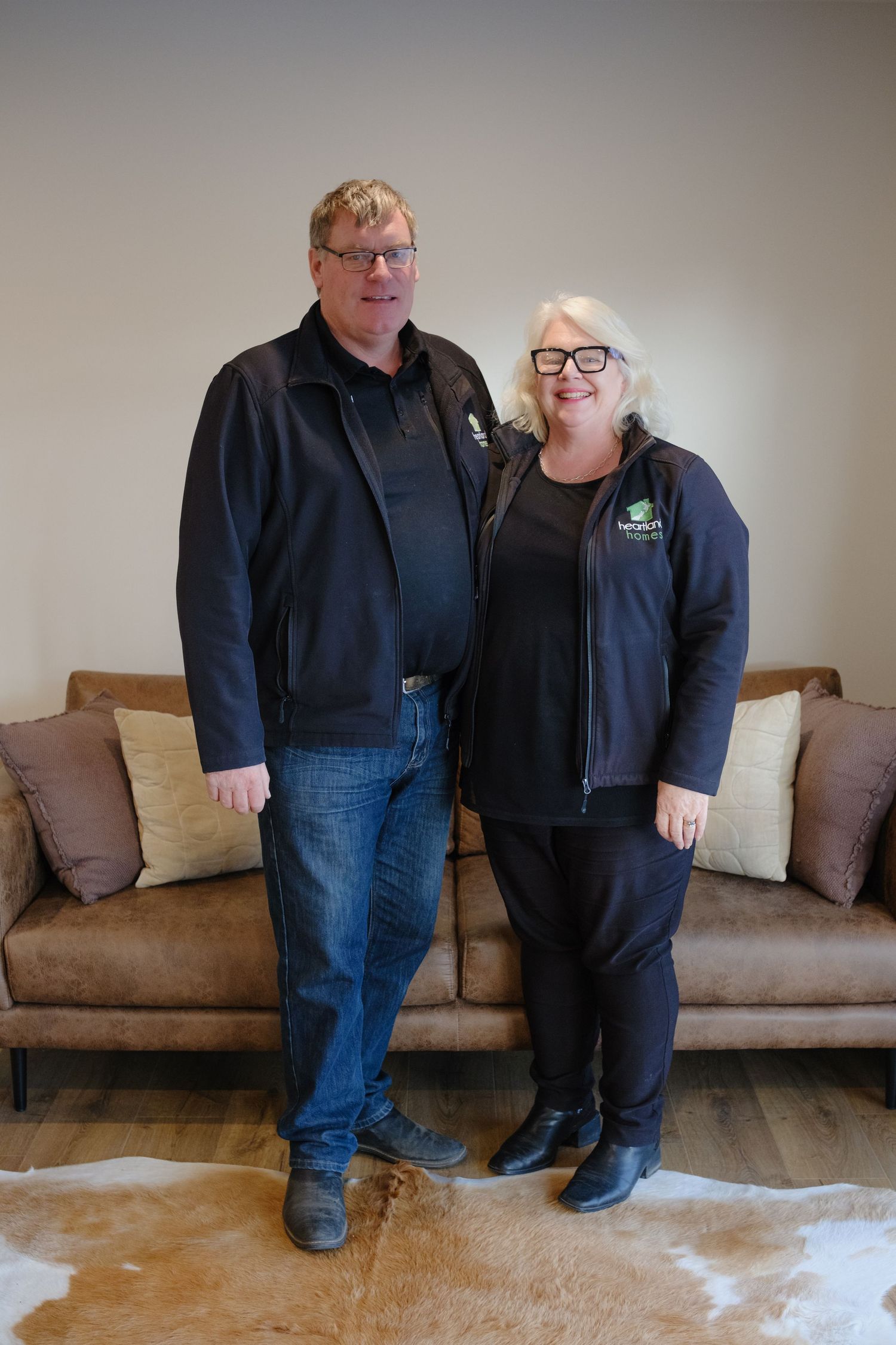Two professionals in matching black company jackets smile together in a modern living room with a leather sofa.