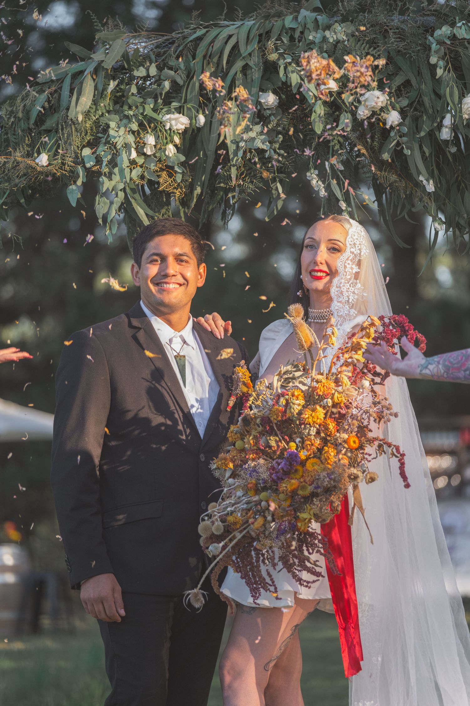 Smiling bride and groom stand under floral arch with confetti, bride holding colorful dried flower bouquet with red ribbon.