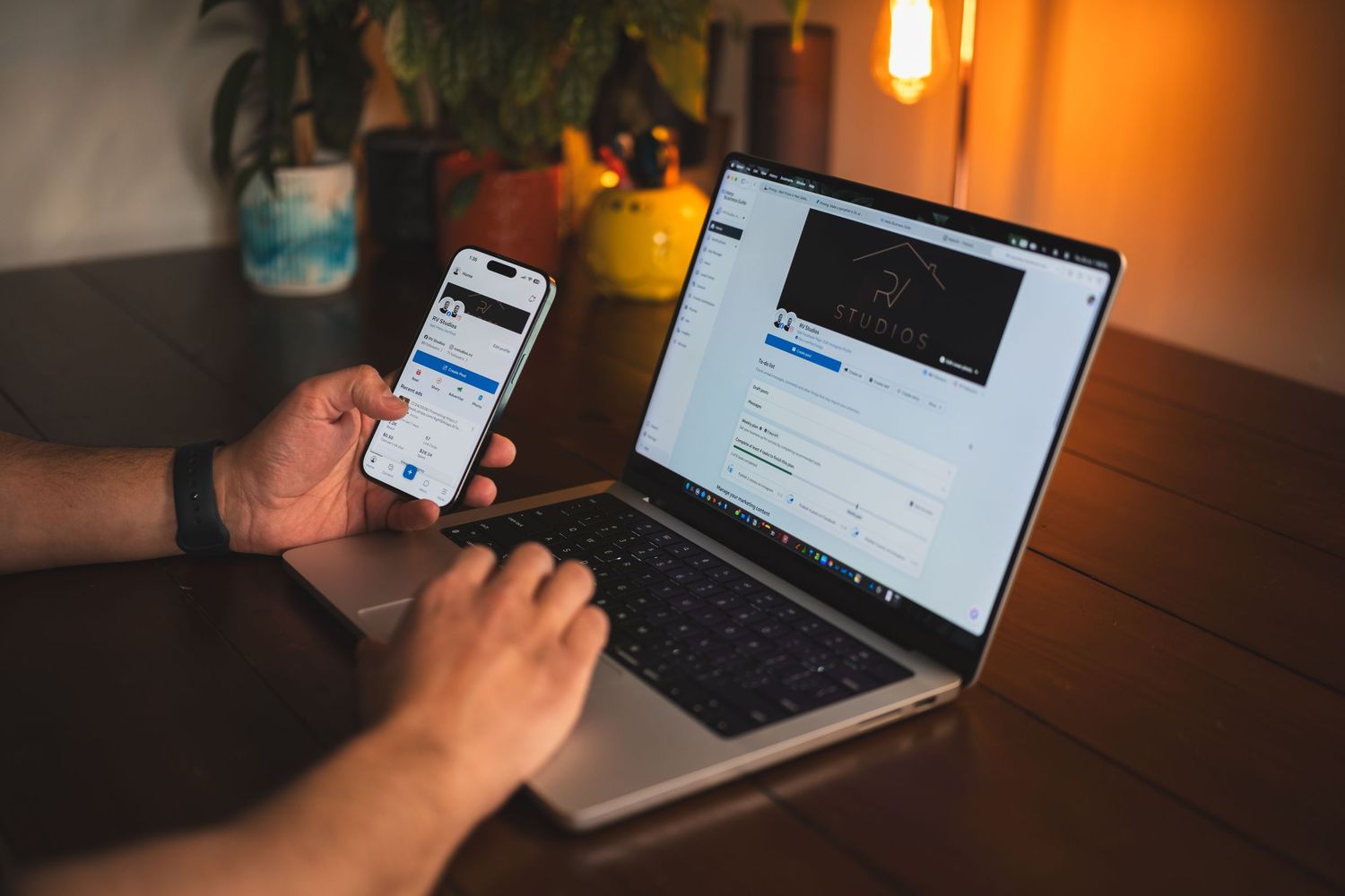 Person using smartphone and laptop simultaneously in a cozy, warmly lit home office setup.