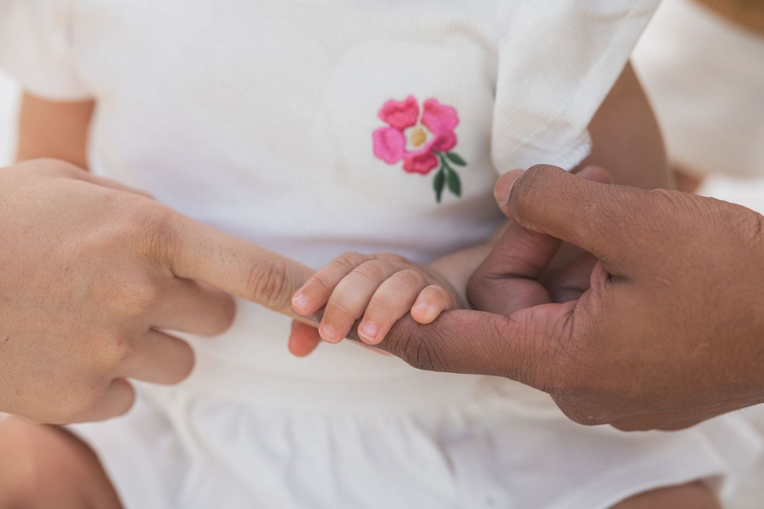 Close-up of a baby holding an adult's finger, wearing a white outfit with a pink flower embroidery.