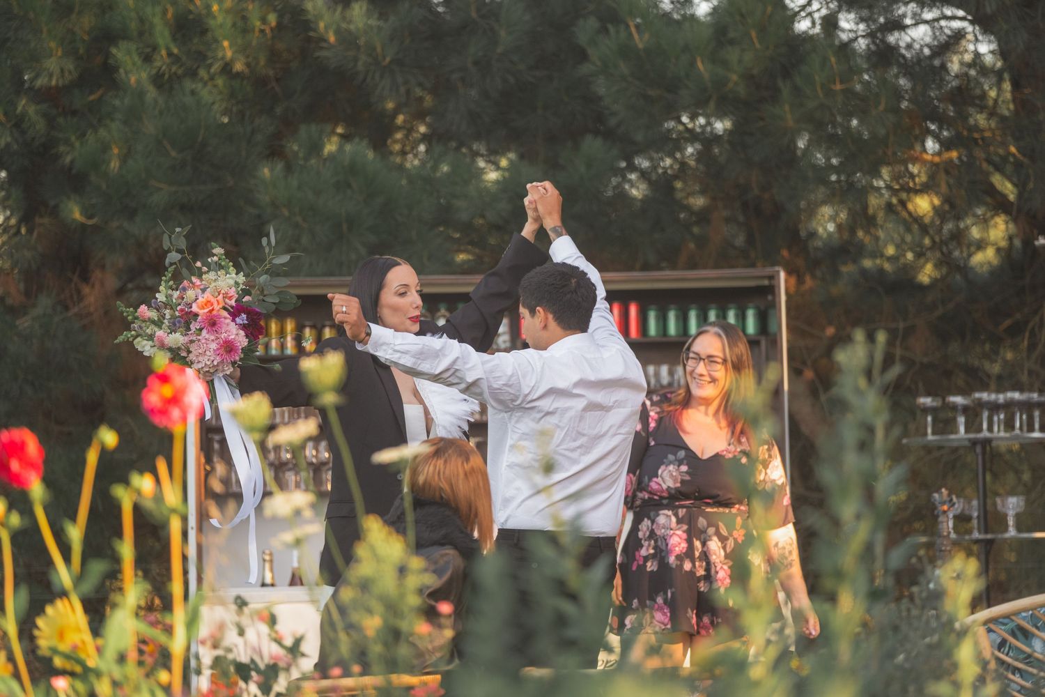 Couple dancing outdoors at a garden party with a bar setup and colorful flowers in the background.