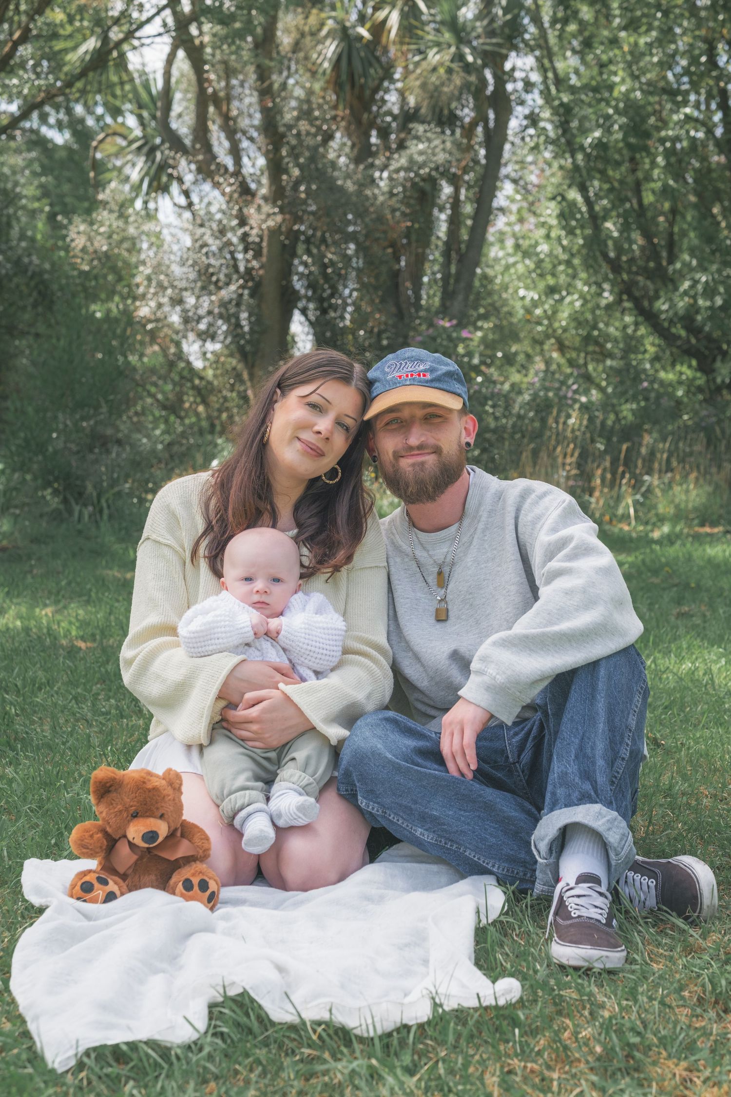 Young family sitting on white blanket in park, mother holding baby with teddy bear, father in cap beside them.