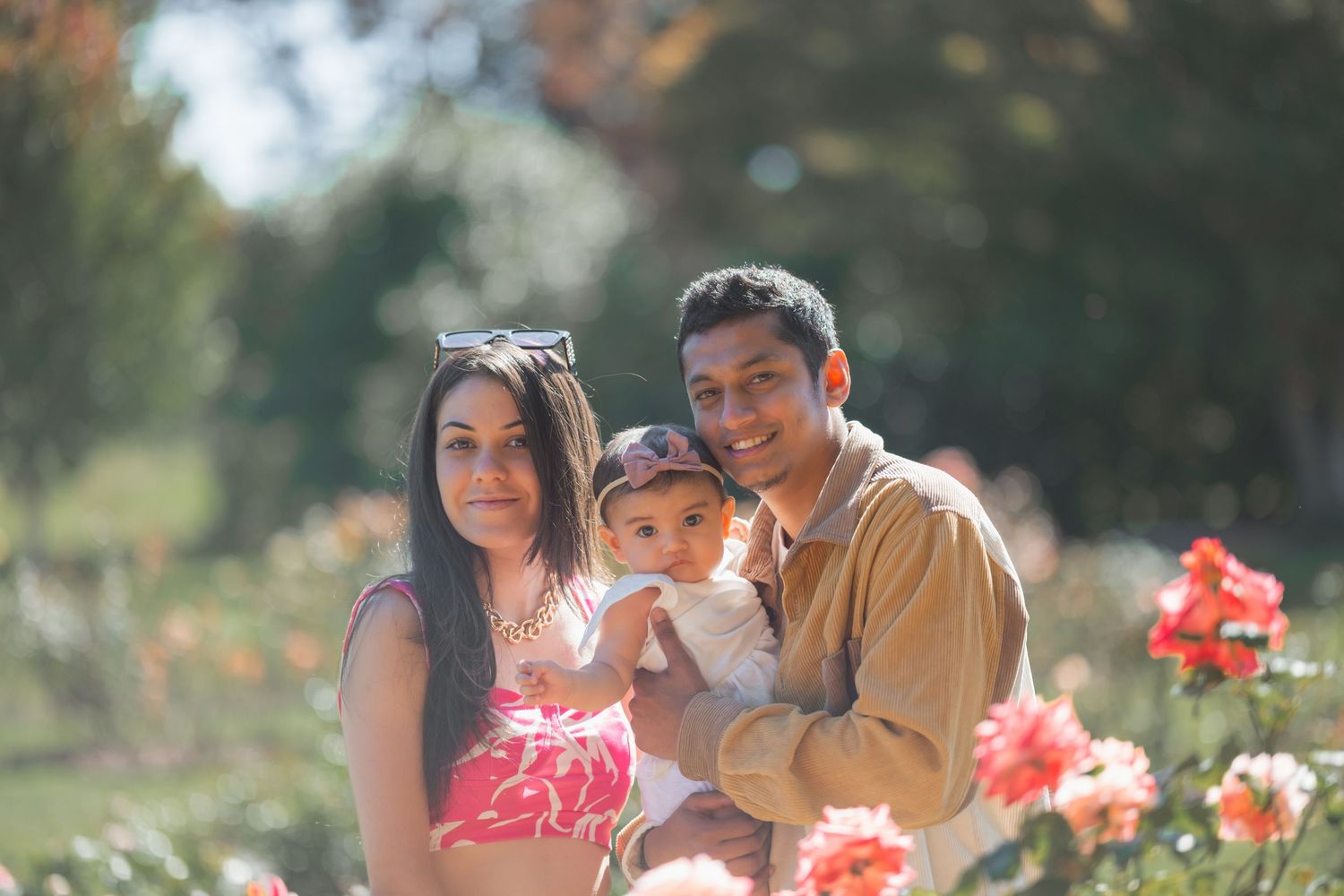 A family poses together in a garden with red flowers blooming in the foreground on a sunny afternoon.