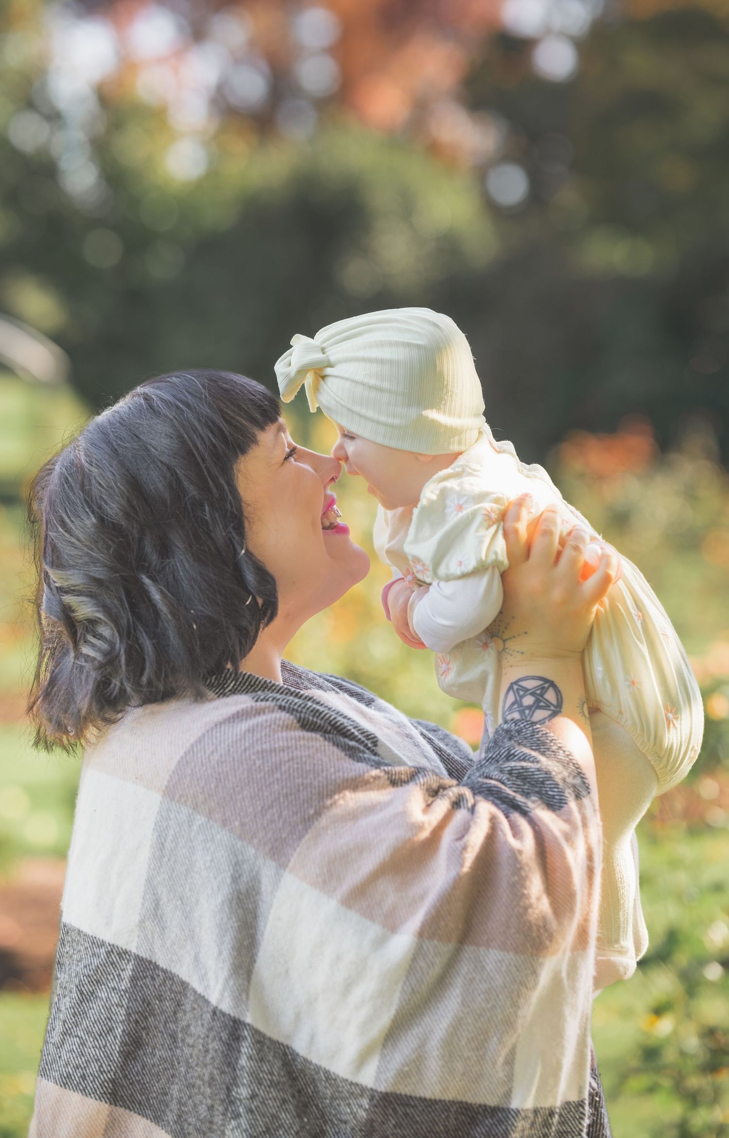 A parent in a striped sweater lifts and cuddles a baby wearing a cream-colored bonnet in an autumn outdoor setting.