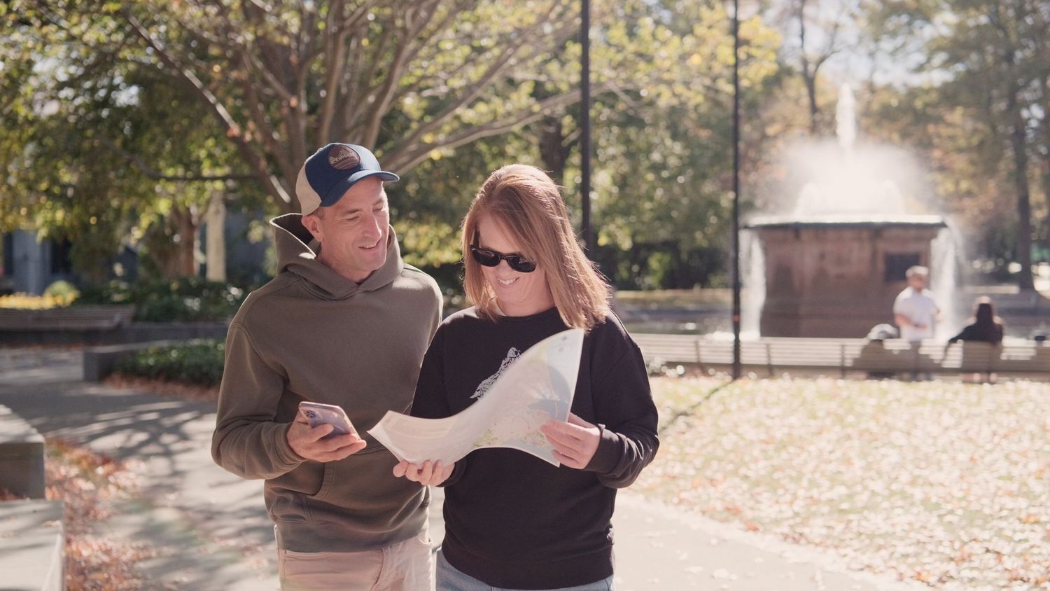 Two people reviewing a map and phone outdoors in a sunny park with a fountain in the background.