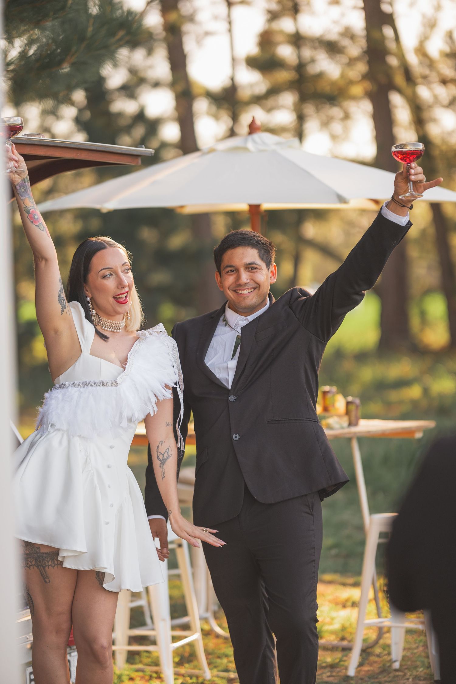 A joyful couple in wedding attire raises glasses outdoors, woman in white mini dress, man in black suit, celebrating.