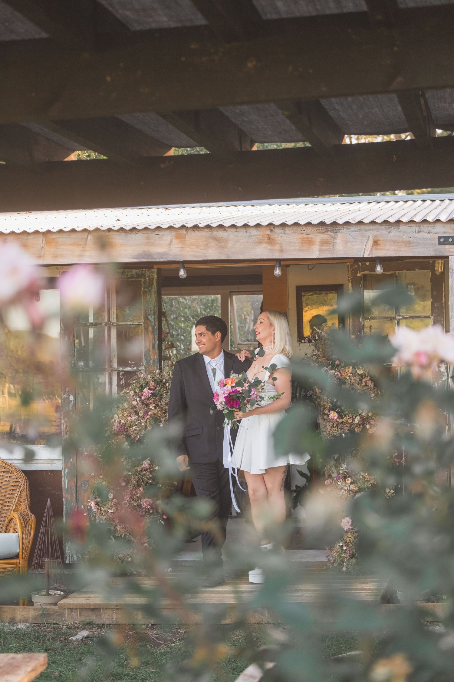 Couple in wedding attire standing on a rustic wooden porch surrounded by blooming roses and floral wreaths.