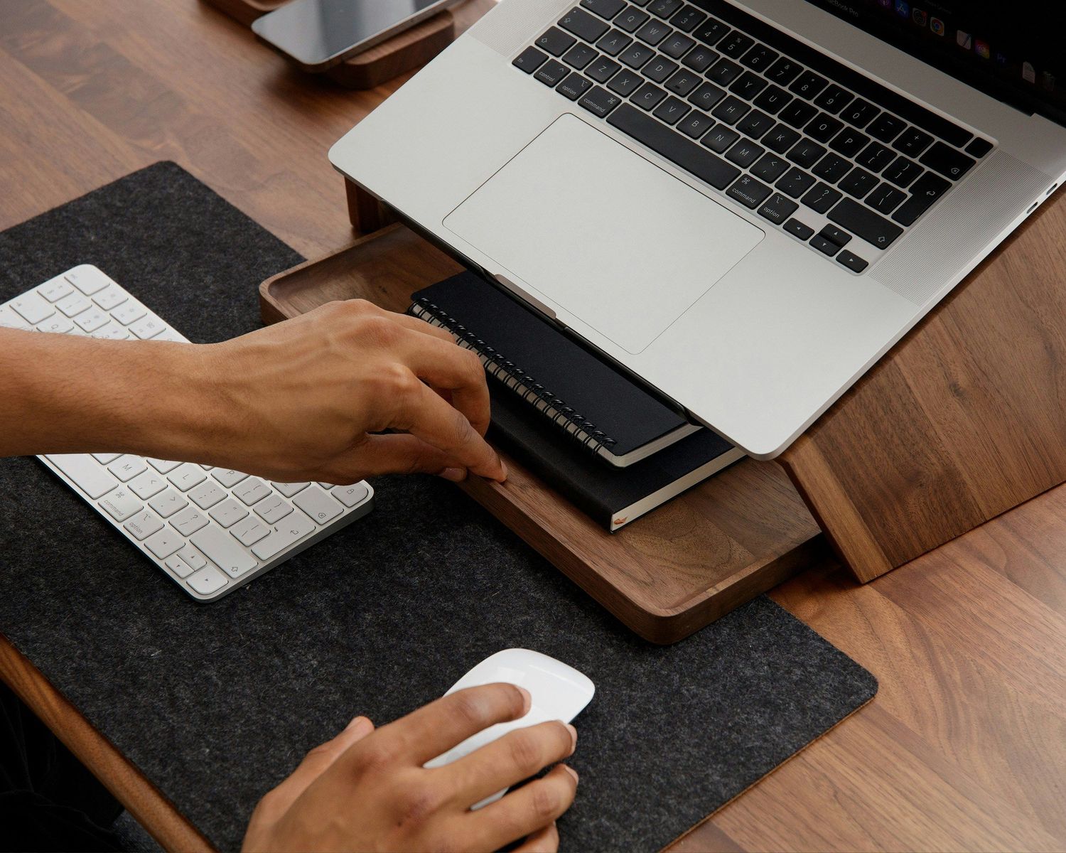 Person using a Magic Mouse on a felt desk pad beside a wooden laptop stand with MacBook and keyboard.