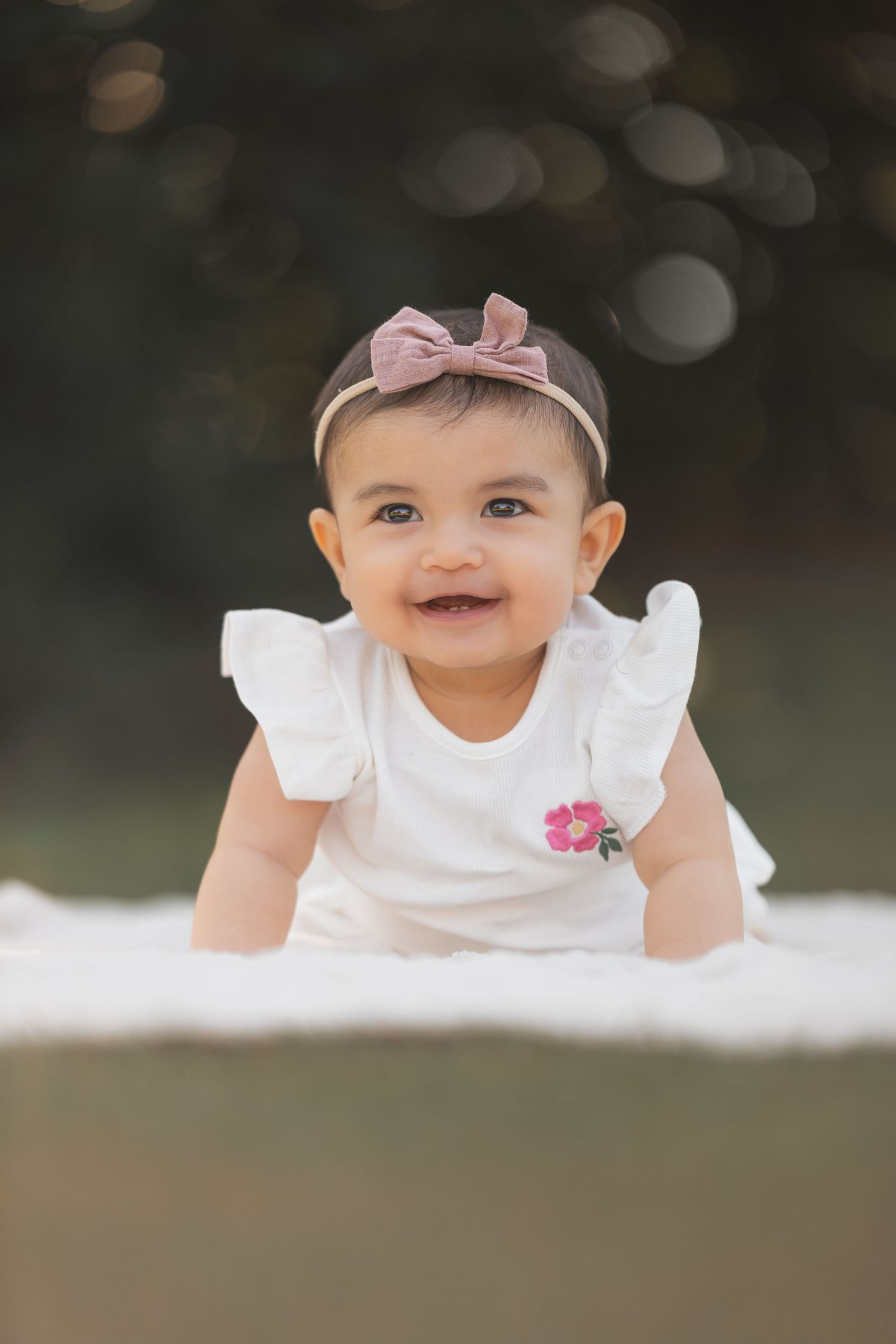 A baby in a white dress with pink bow headband smiles while laying on a blanket outdoors.