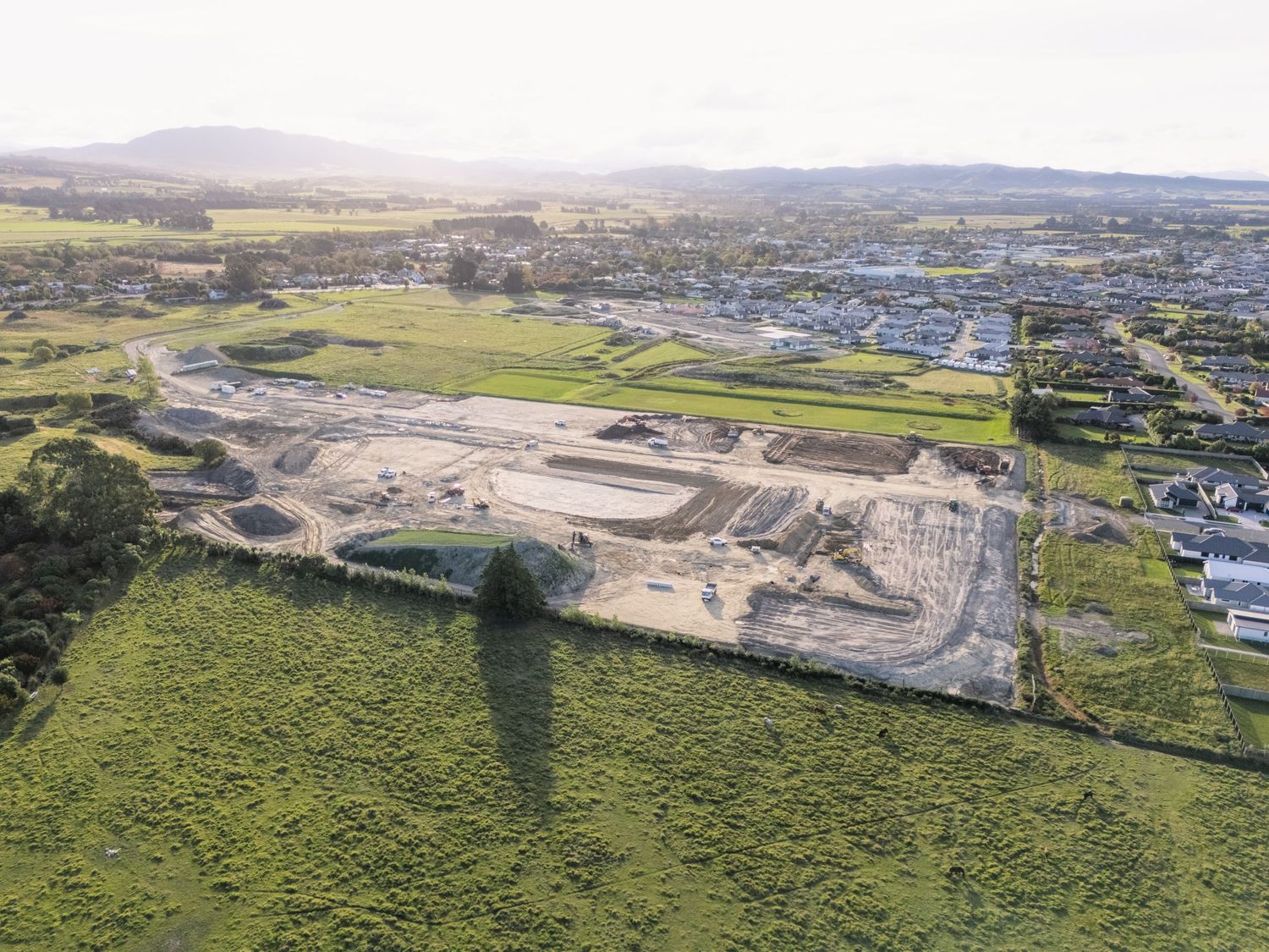 Aerial view of a large construction site development on green farmland with residential suburbs and hills in the background.