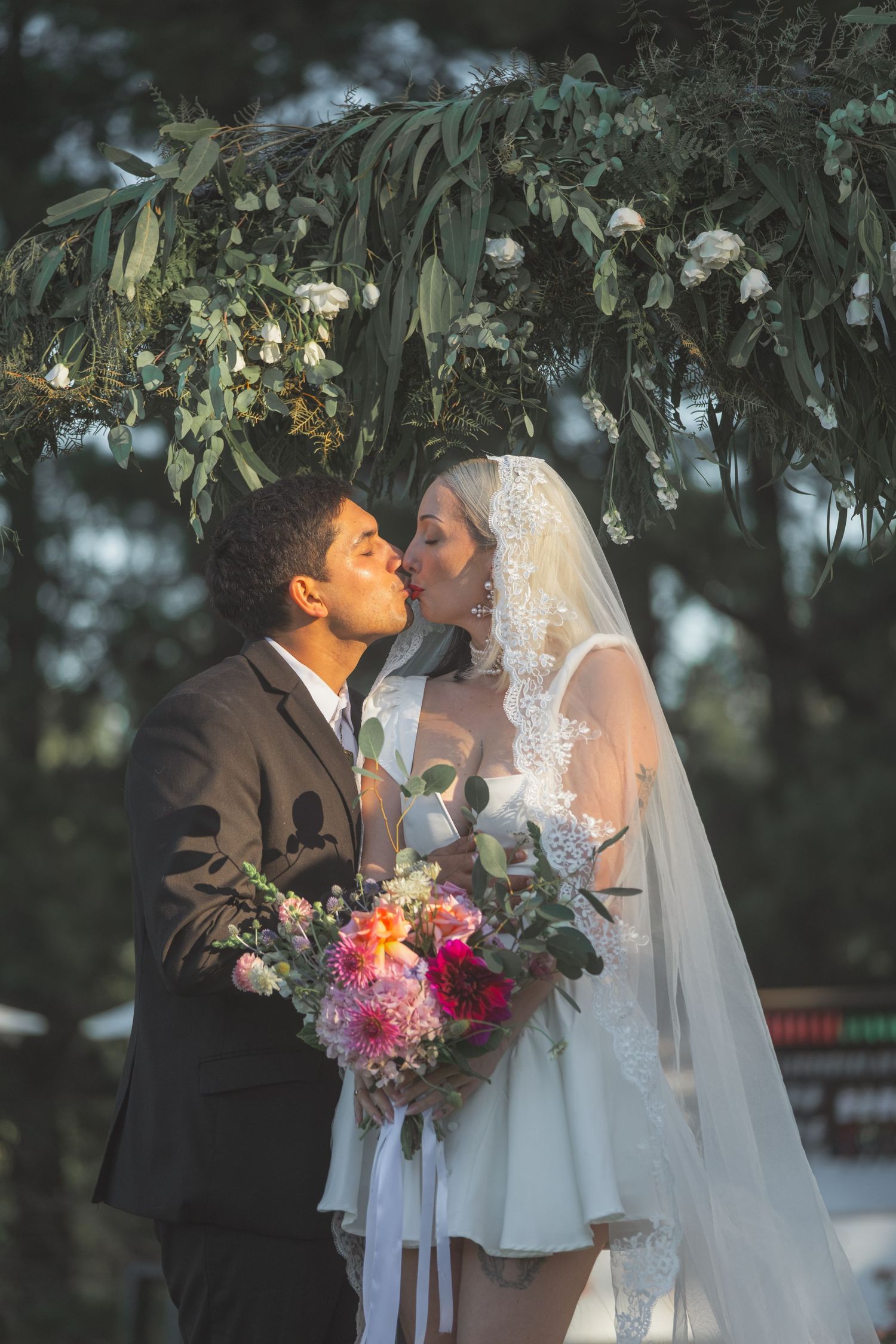 Bride and groom share a kiss under a floral arch, bride holding a colorful bouquet in an outdoor wedding ceremony.