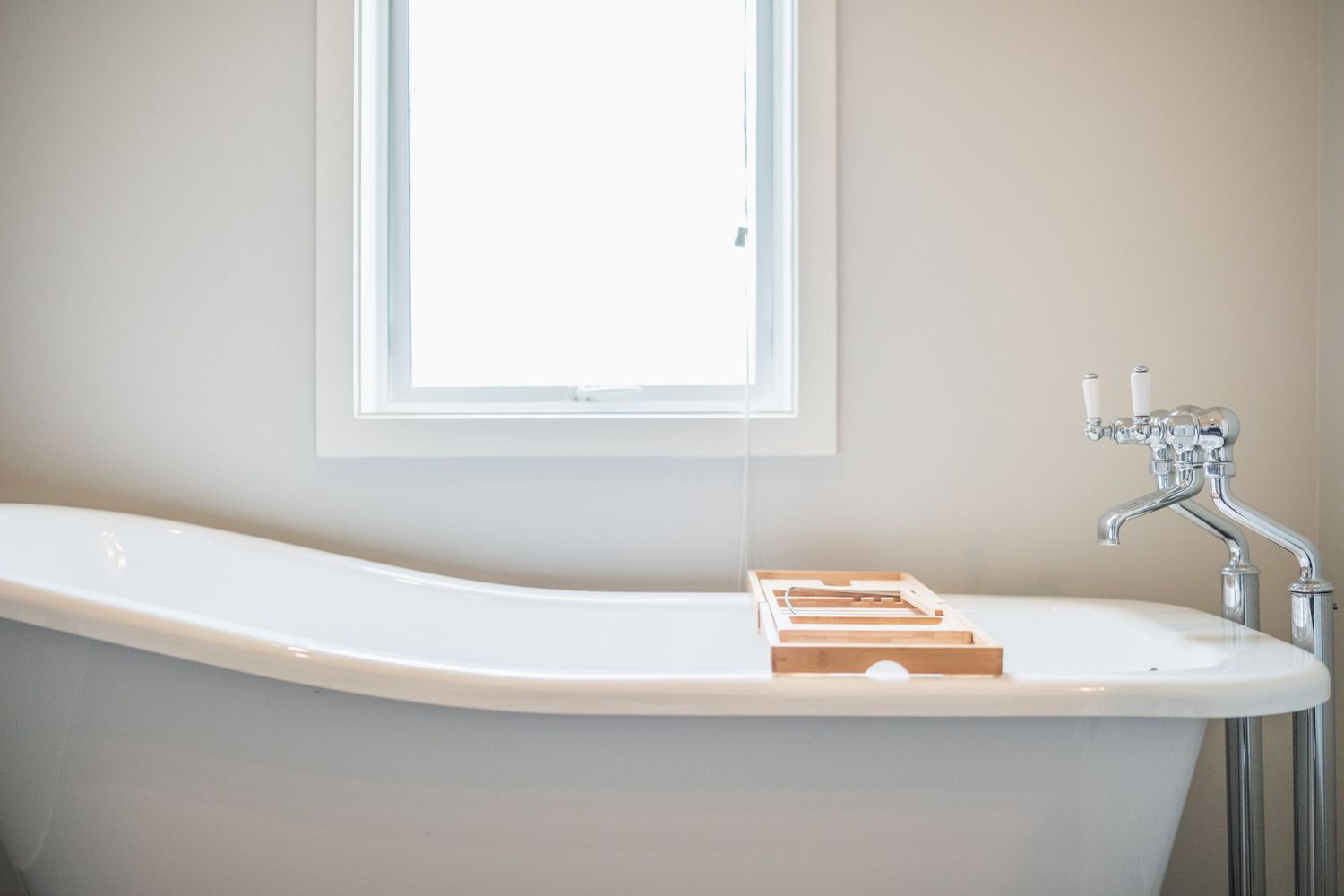 White freestanding bathtub with wooden tray near bright window in modern minimalist bathroom.