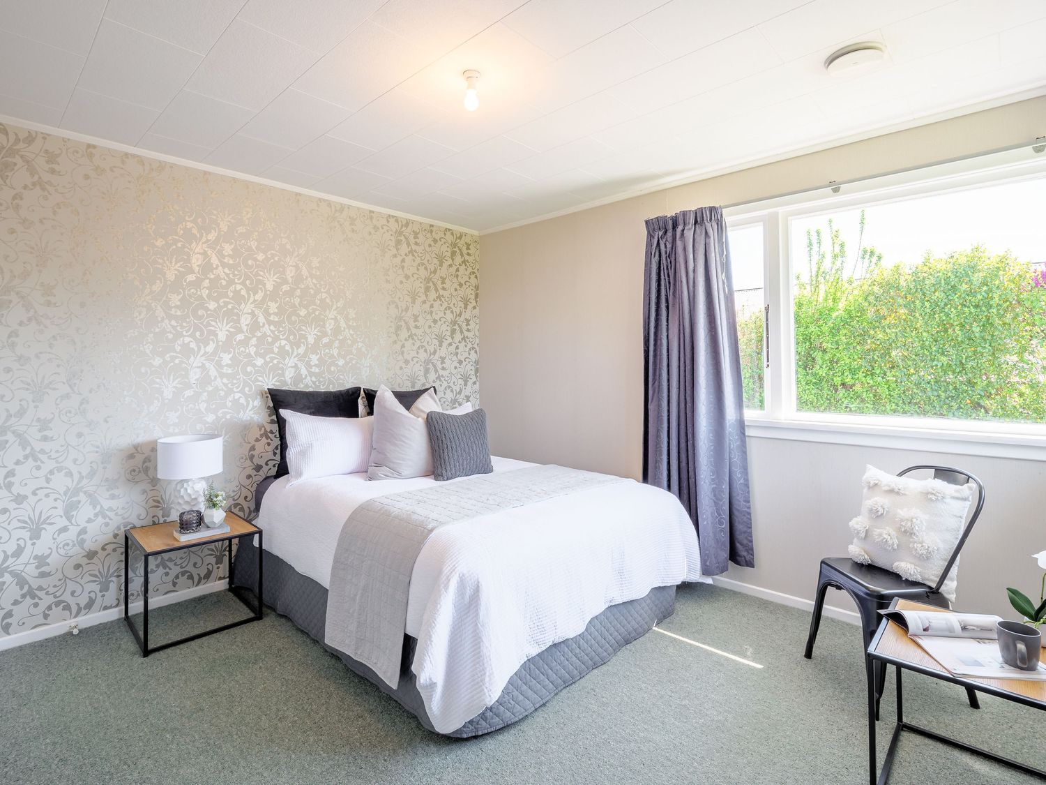 Modern bedroom with white bedding, silver wallpaper, grey curtains, and a large window overlooking greenery.