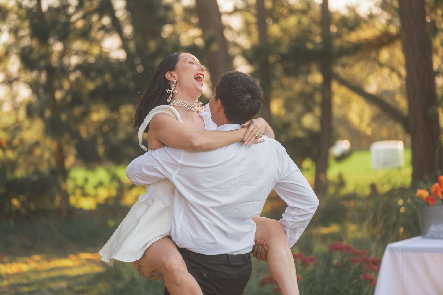 A joyful couple shares a romantic moment outdoors, with the groom lifting his laughing bride in a sunlit garden.
