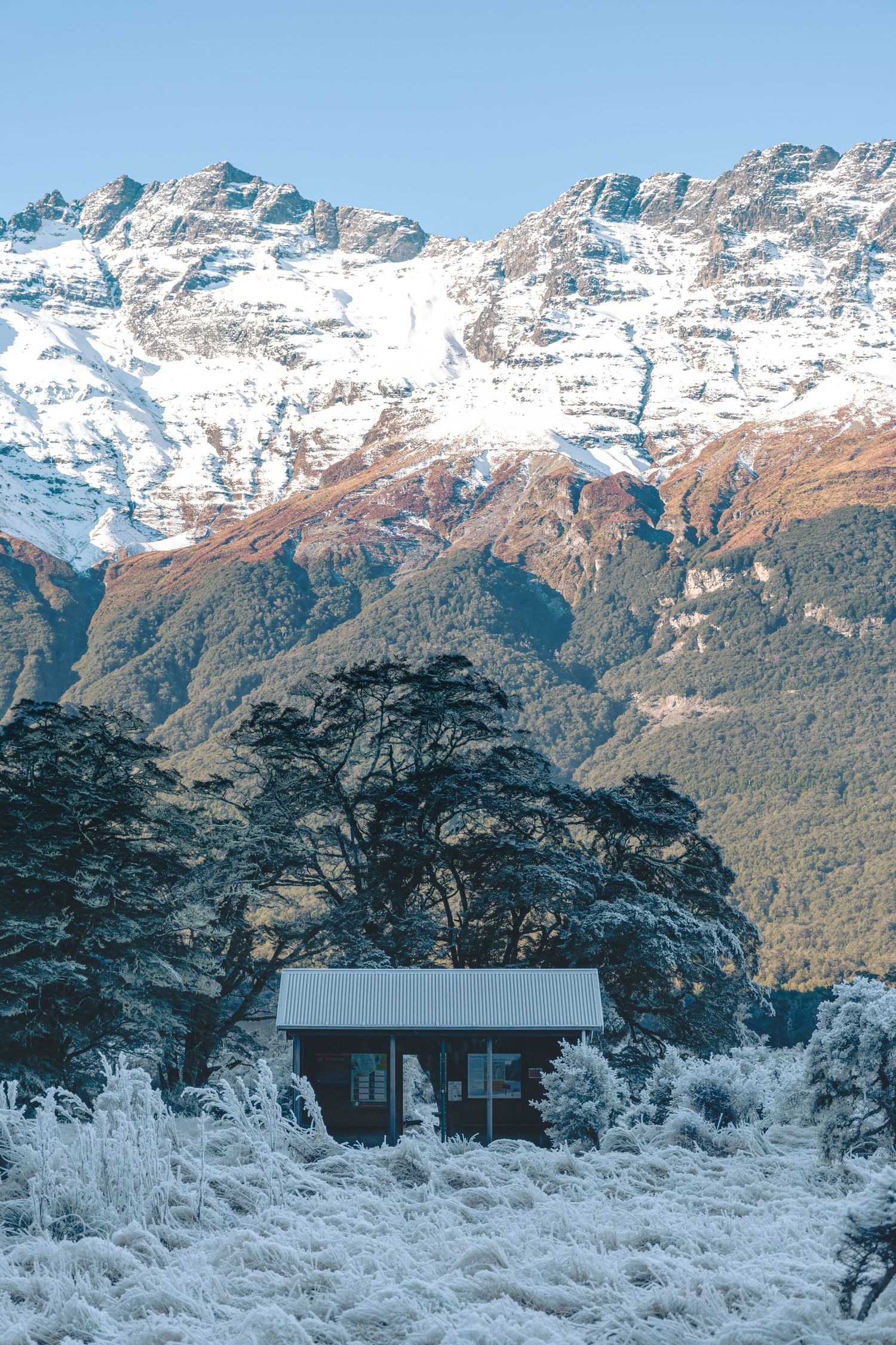 A small cabin nestled in a frosty valley with snow-capped mountains towering in the background against a blue sky.