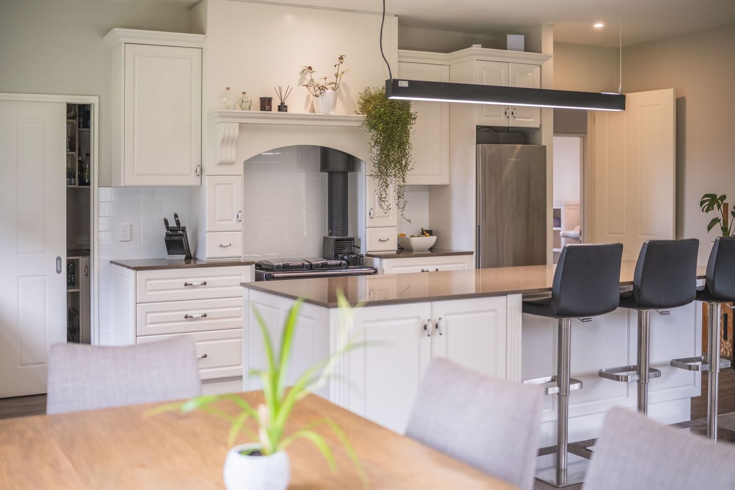 Modern white kitchen with island, bar stools, stainless steel appliances, and pendant light over wooden dining table.