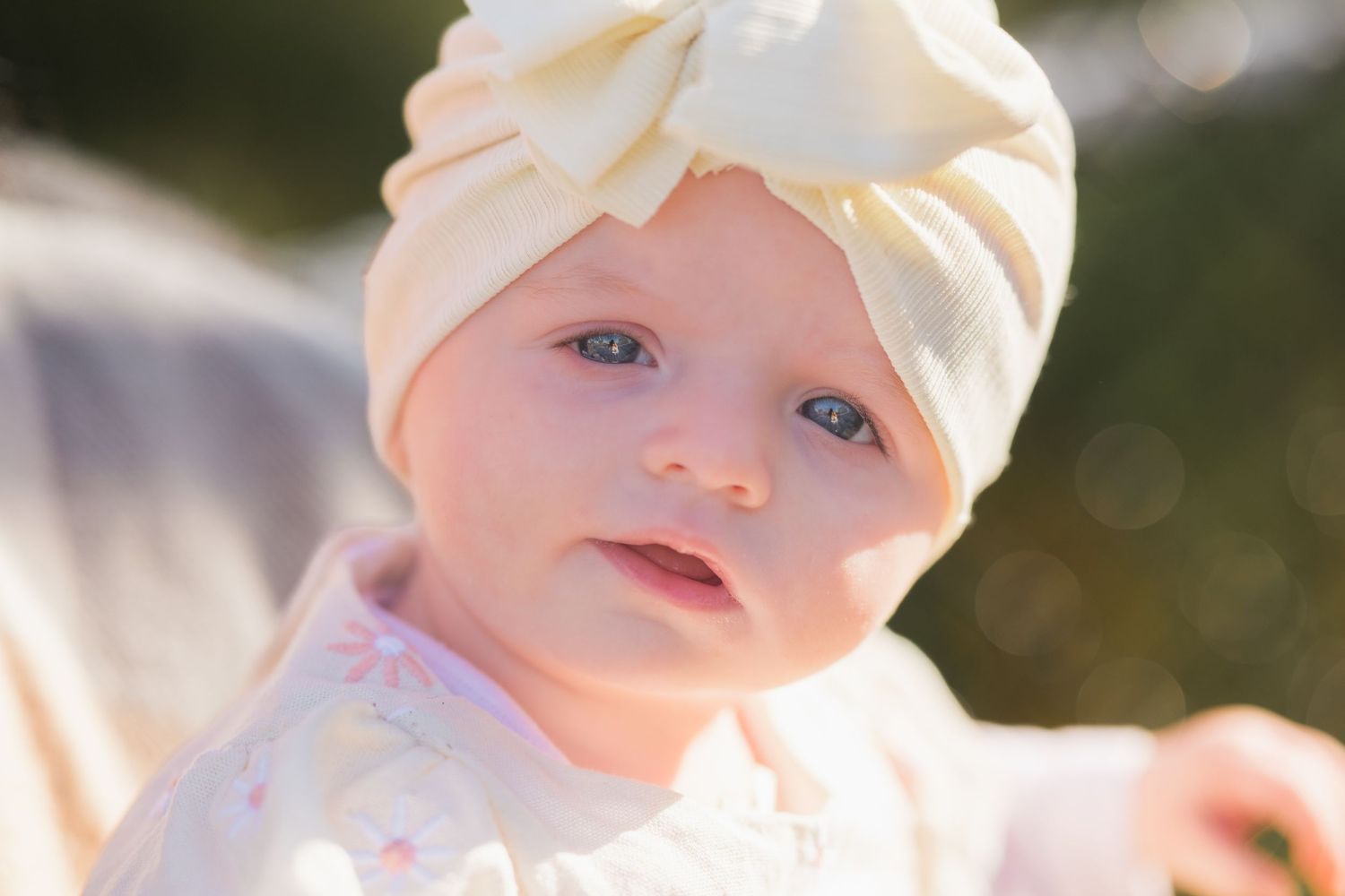 A baby wearing a cream turban-style headband sits outdoors in soft sunlight.