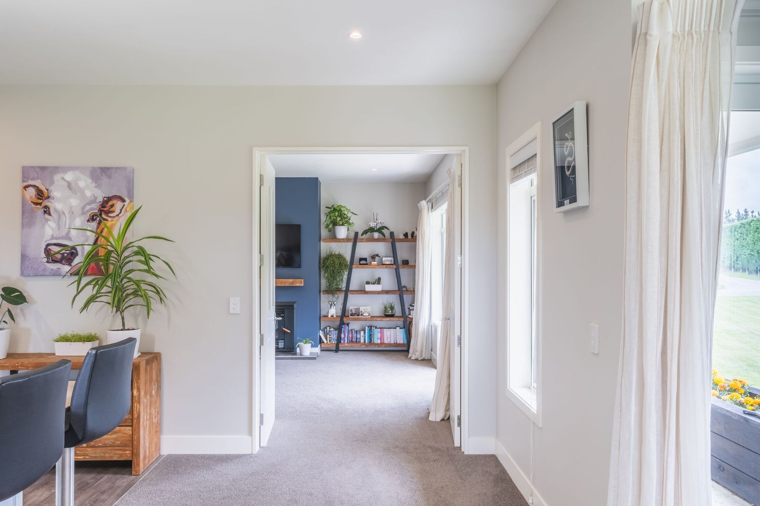 Bright hallway with grey carpet, white walls, a blue dining chair, green plant, and a bookshelf visible through the doorway.