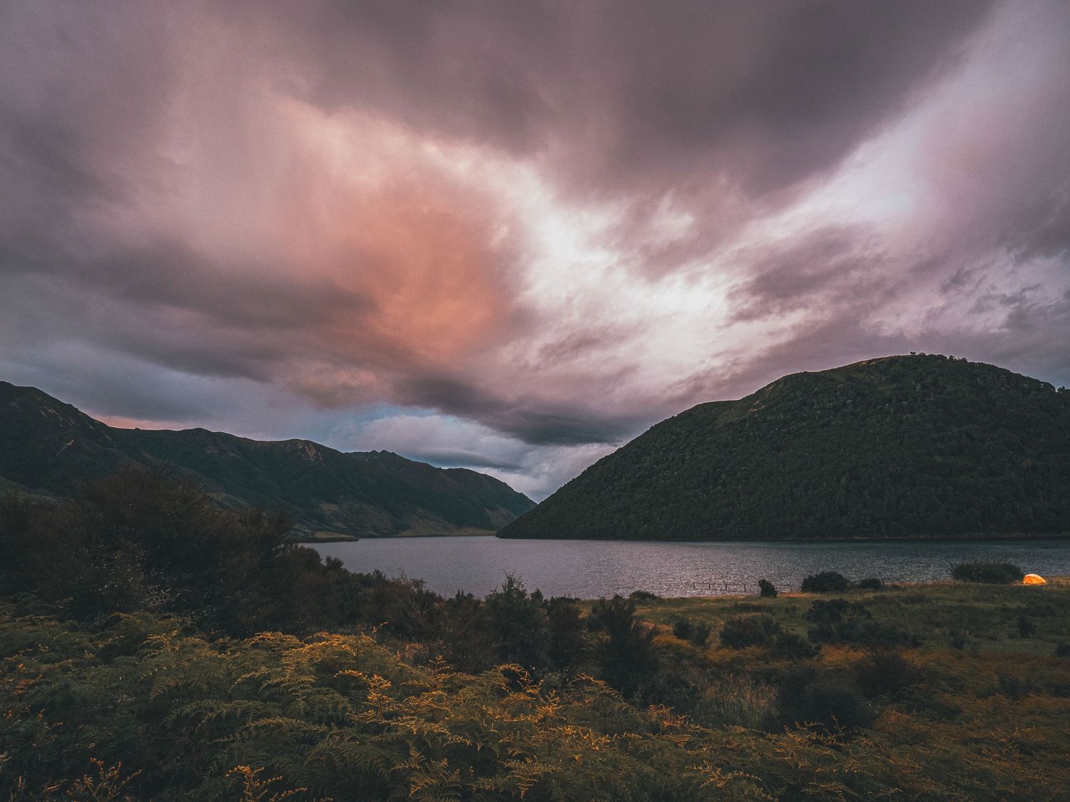 Dramatic pink and purple storm clouds hover over a mountain lake at dusk with glowing lights visible along the shore.