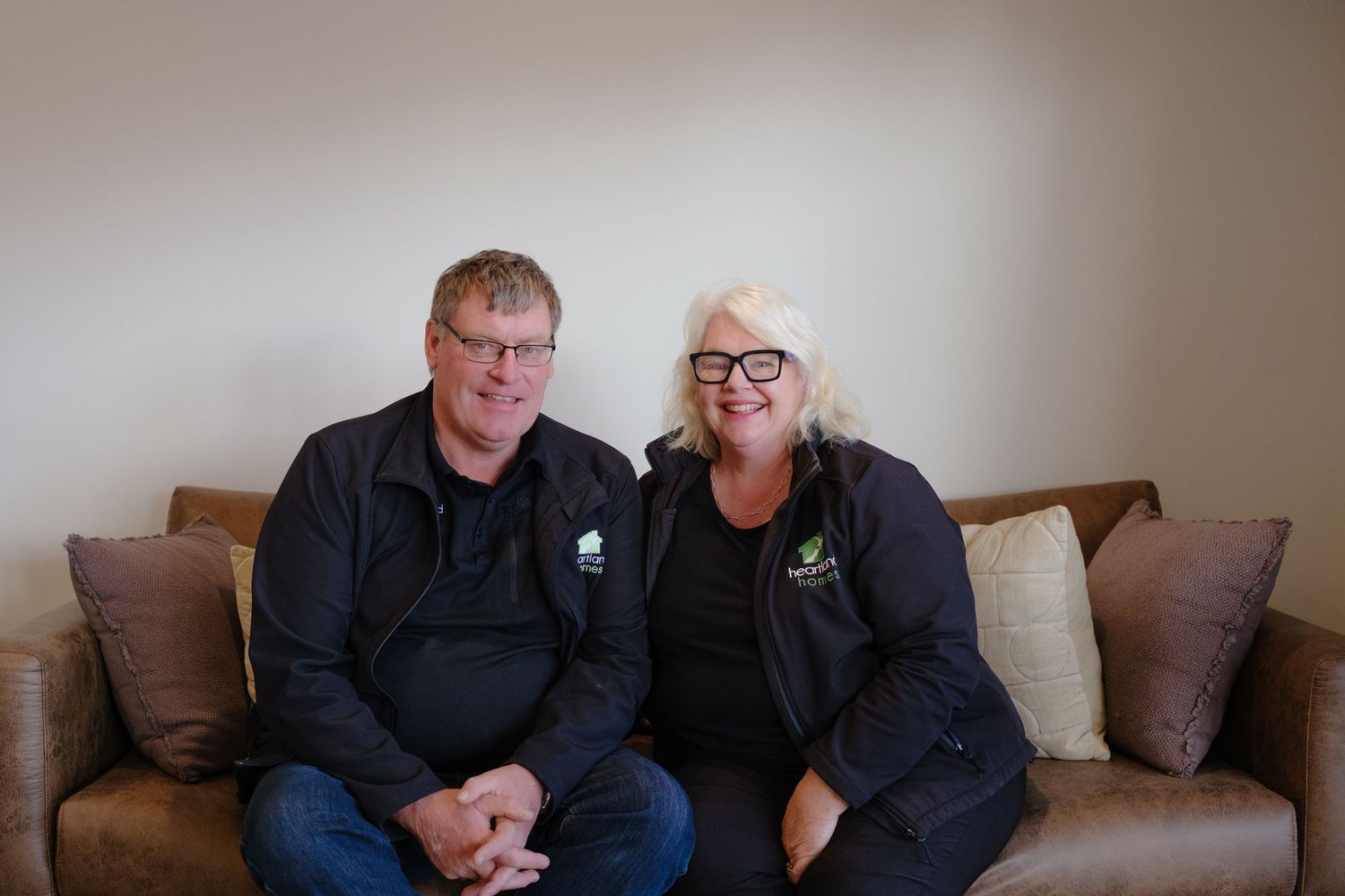 Two smiling adults in matching black jackets sit together on a brown leather couch against a neutral wall.