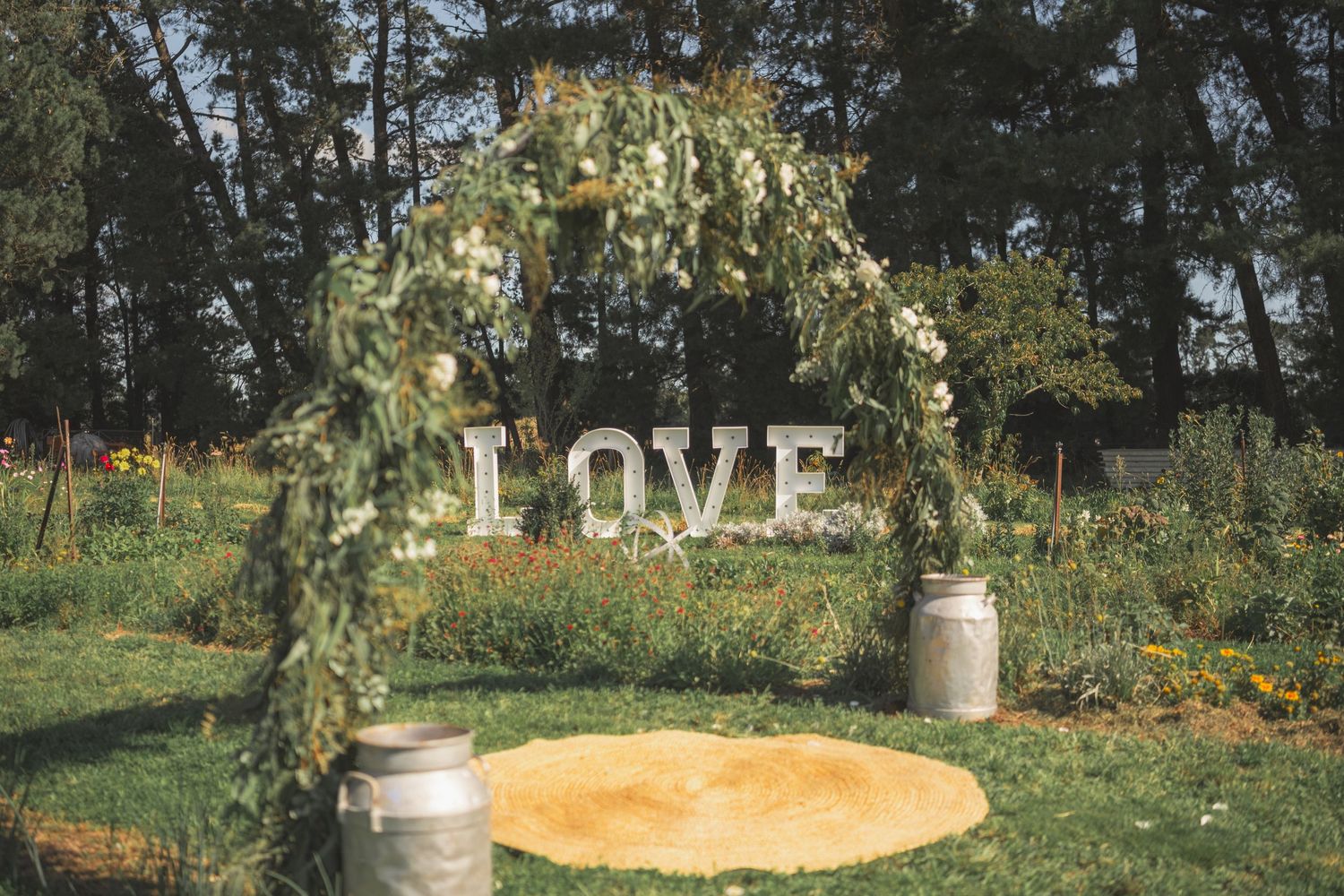 Outdoor wedding arch decorated with greenery and LOVE letters, flanked by rustic milk cans on a golden circle in a garden.