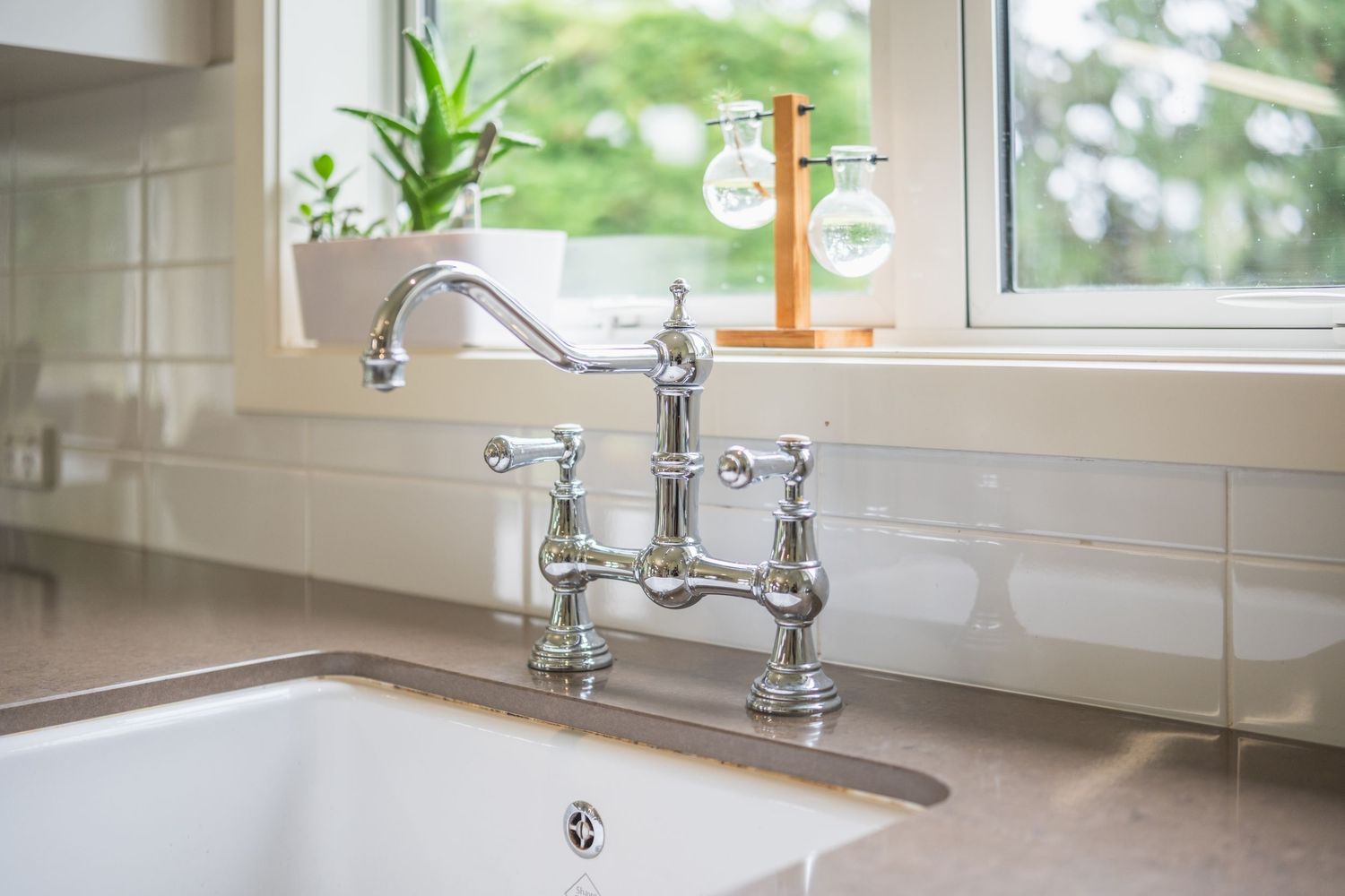 Chrome bridge kitchen faucet over white farmhouse sink with window and green plants in background.