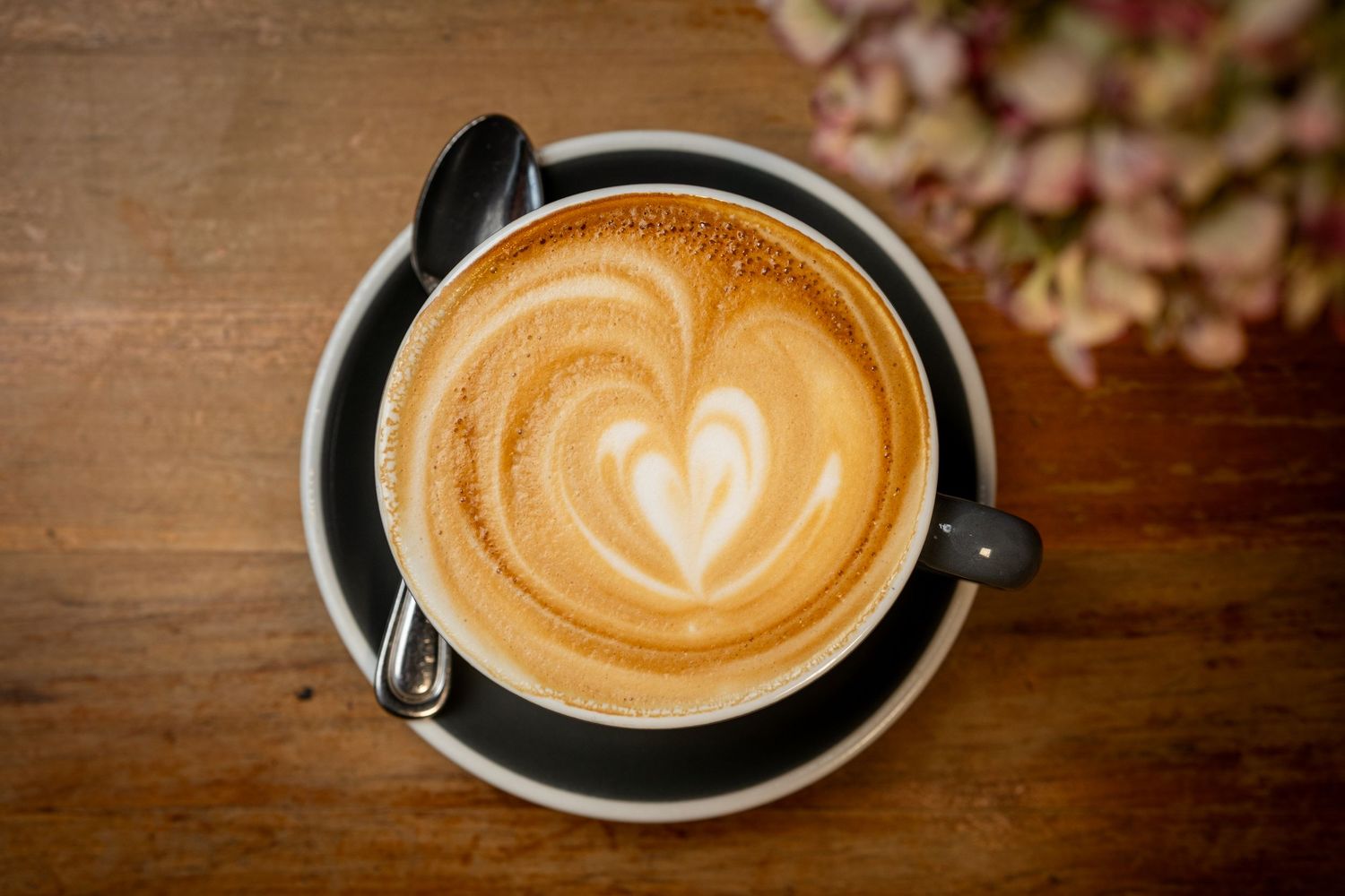 Overhead view of a latte art coffee with heart design in a black cup on saucer, on a wooden table.
