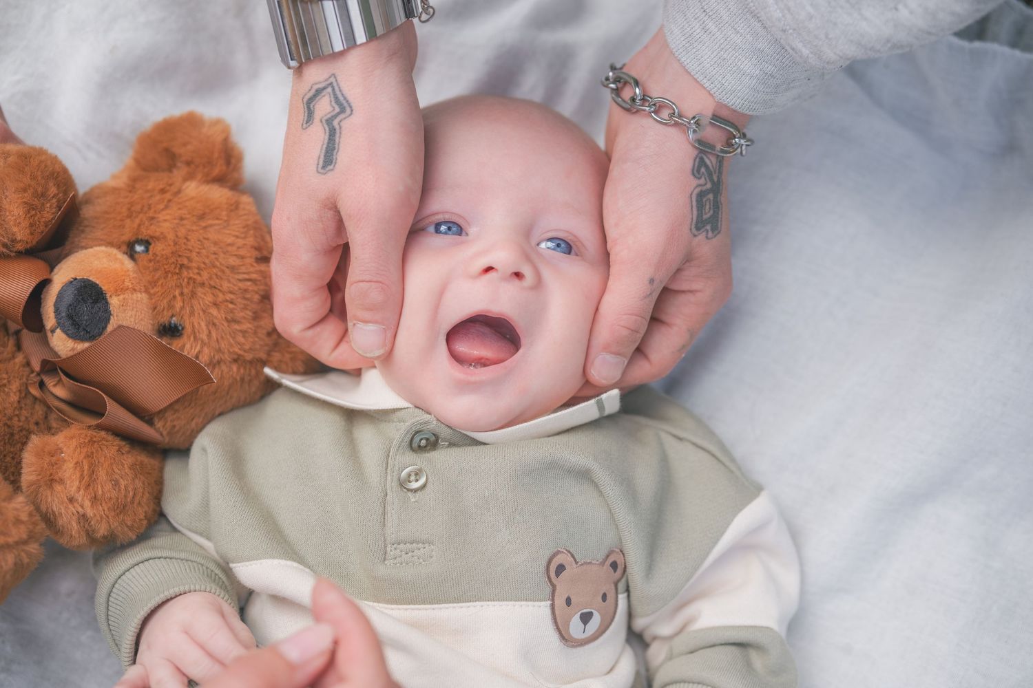 A smiling baby with blue eyes in a bear onesie, held gently by tattooed hands, lying next to a teddy bear.