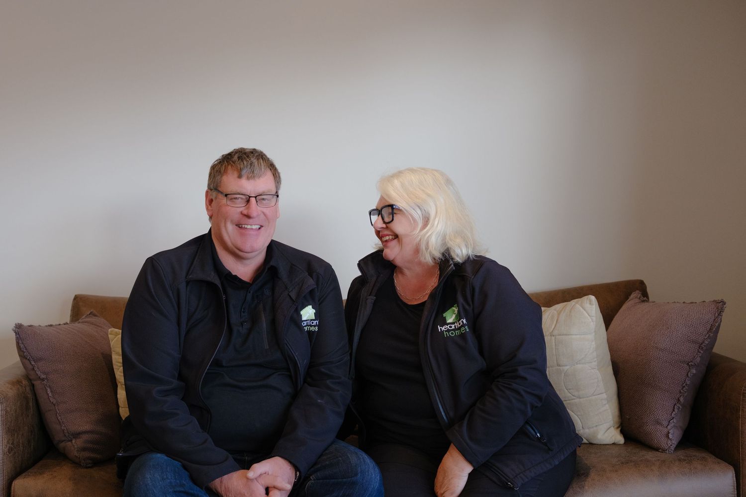 Two smiling adults in matching black jackets sit together on a brown couch in a bright, modern living room.
