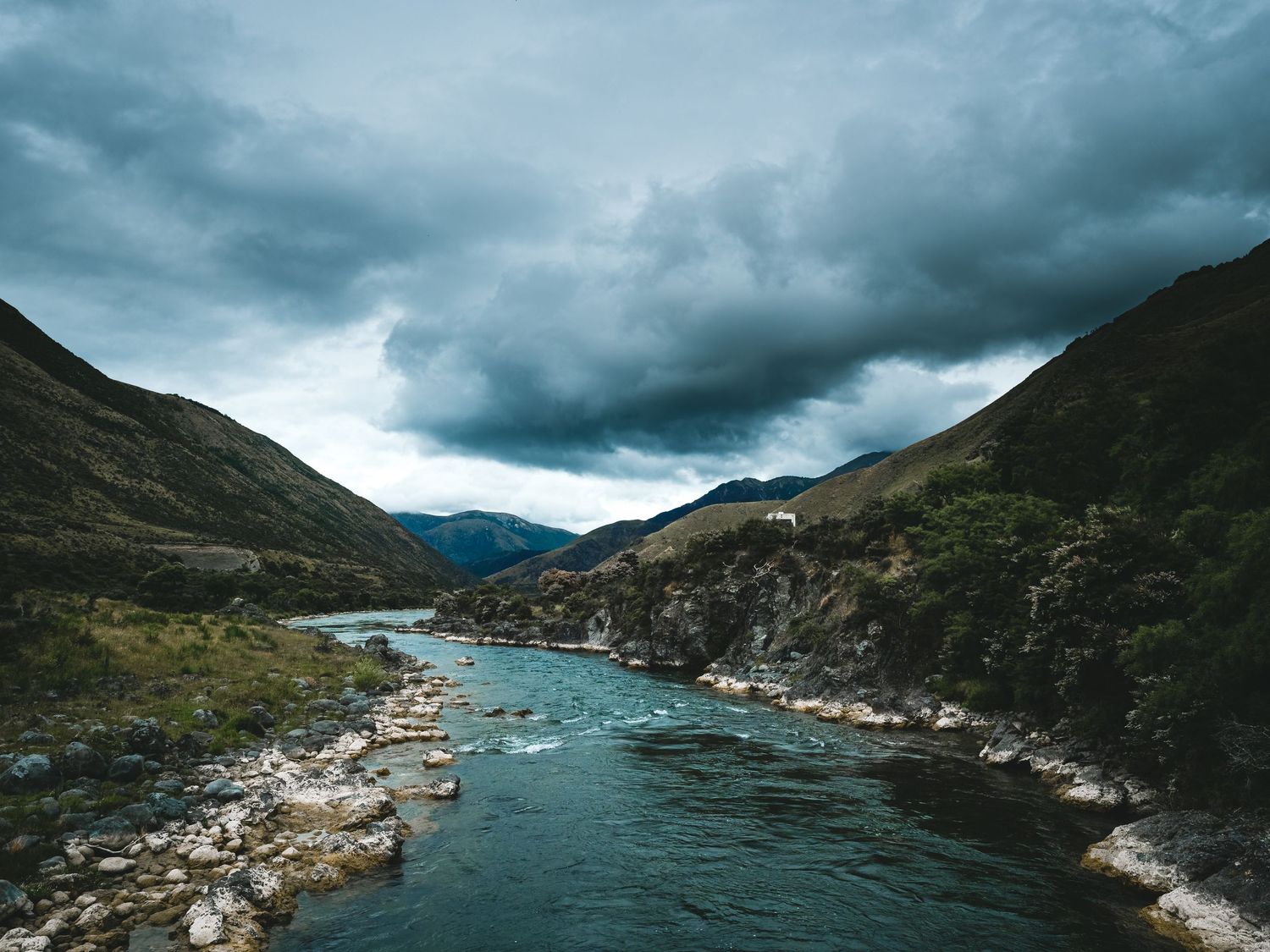 Dramatic panorama of a river nestled between mountains with rocks along either side dotting the riverbank under moody skies.