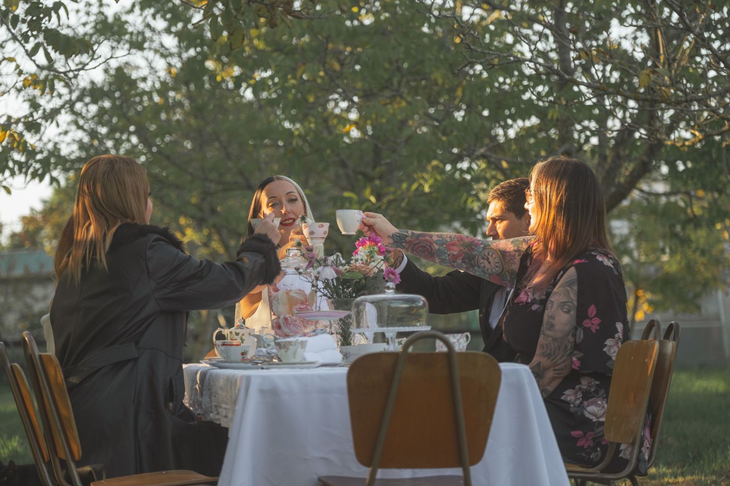 Four women toasting with glasses at an outdoor garden party table with white tablecloth and trees in background.