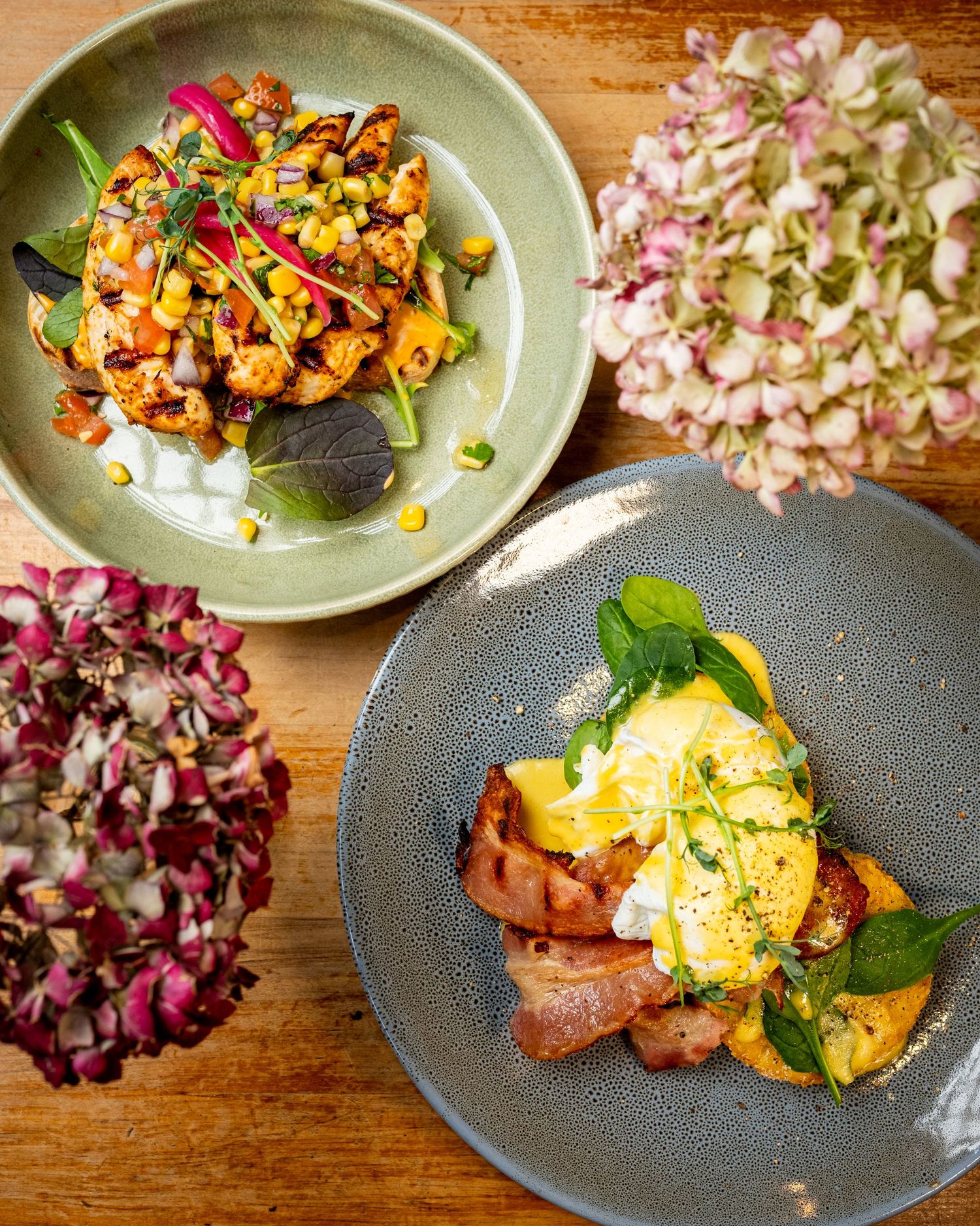 Two gourmet brunch dishes with eggs benedict and roasted vegetables on rustic plates, garnished with fresh herbs and flowers.