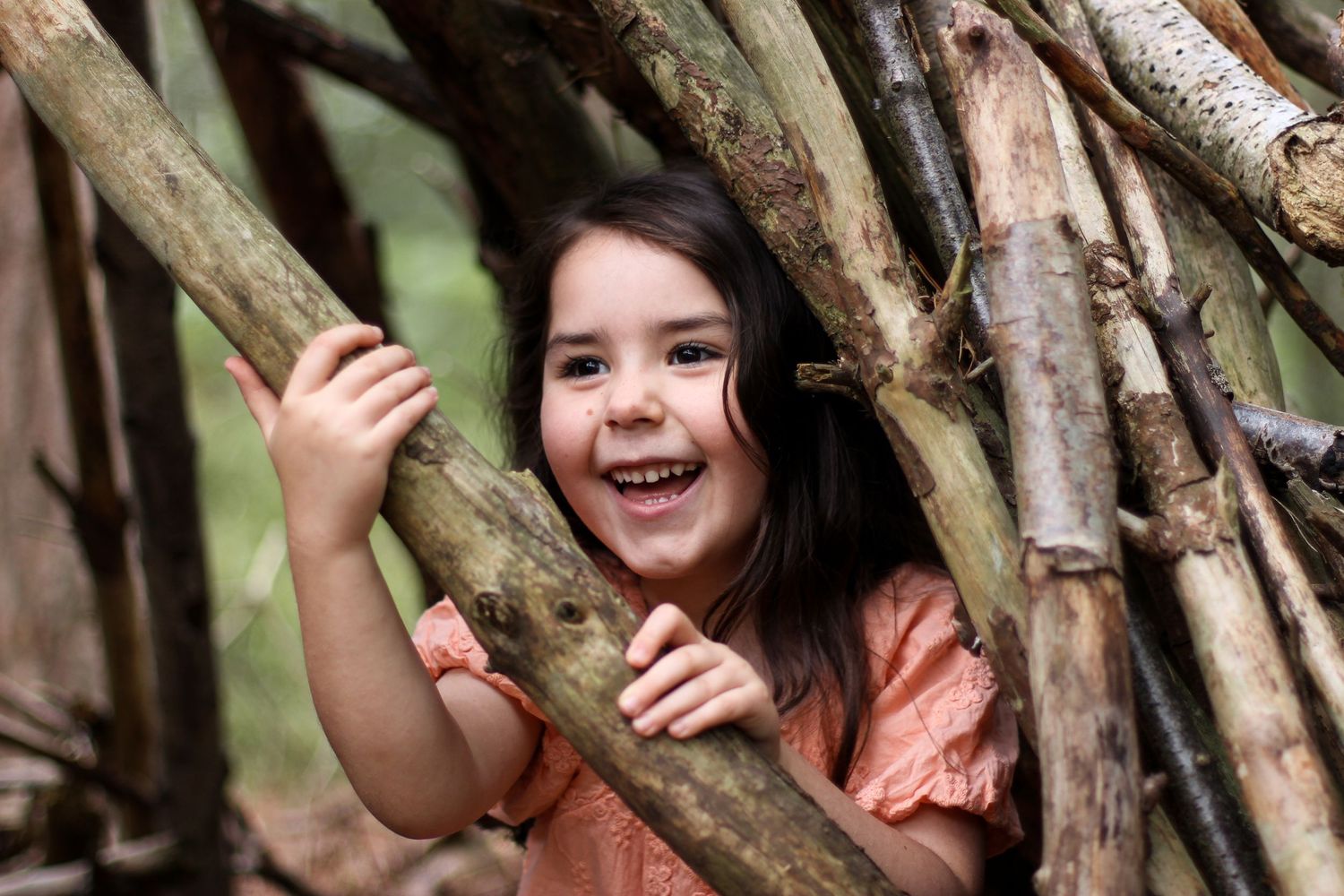 A smiling child peeks through tree branches in the forest while wearing a peach colored top.