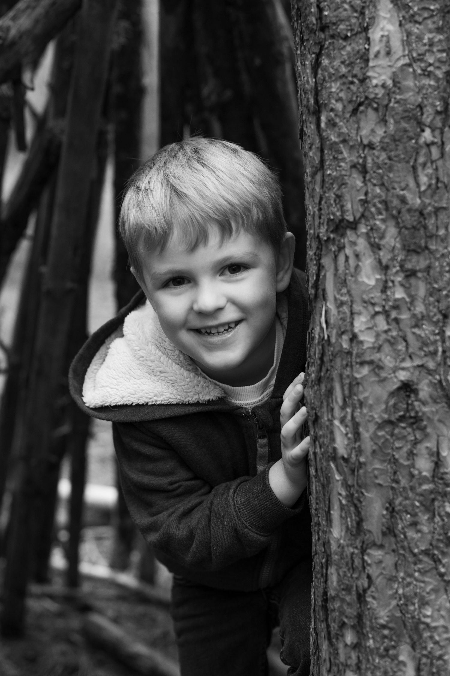 Young child playfully peeks out from behind tree trunks in a black and white forest scene.