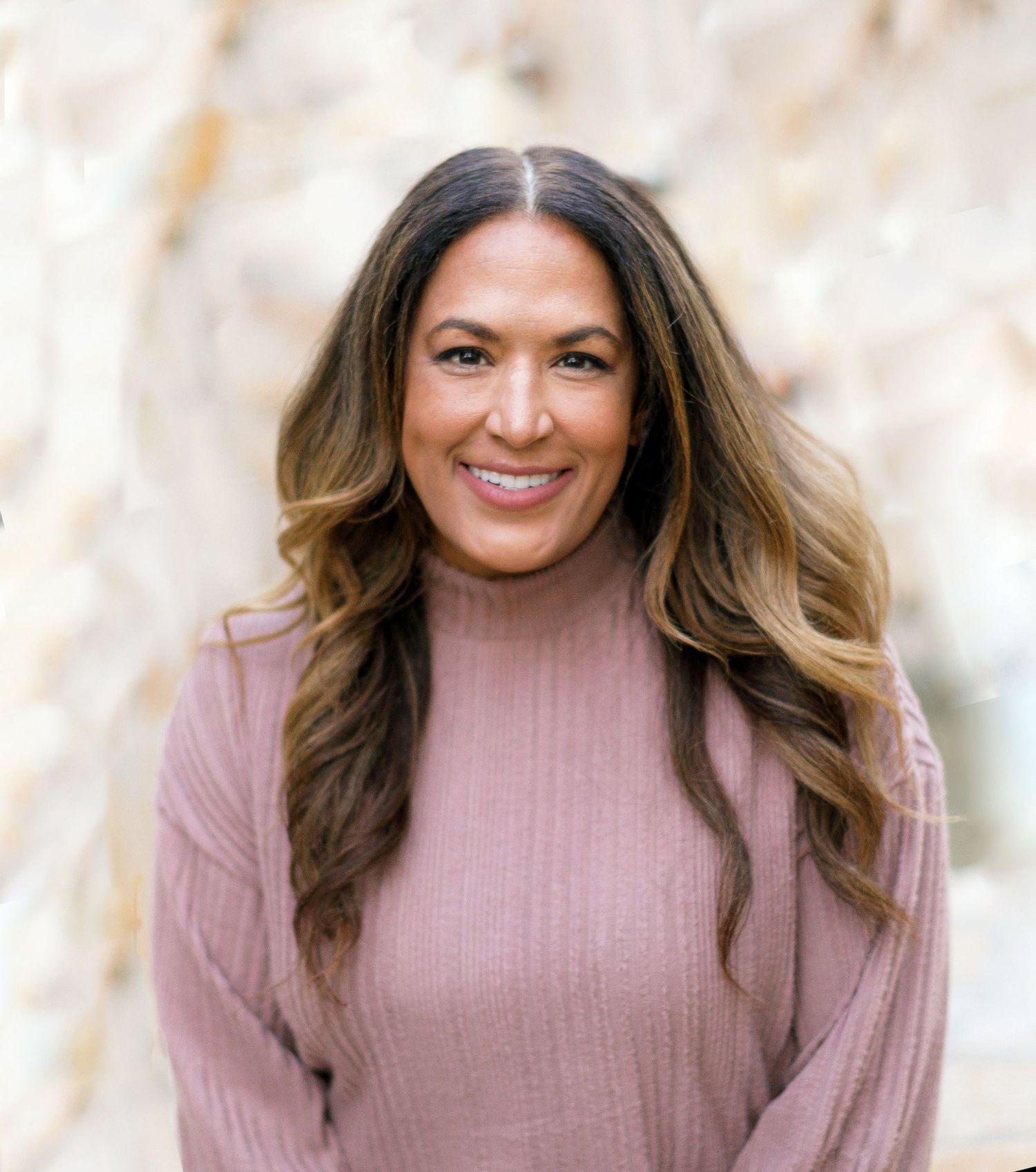 Smiling woman with long wavy brunette hair wearing a pink turtleneck sweater outdoors.