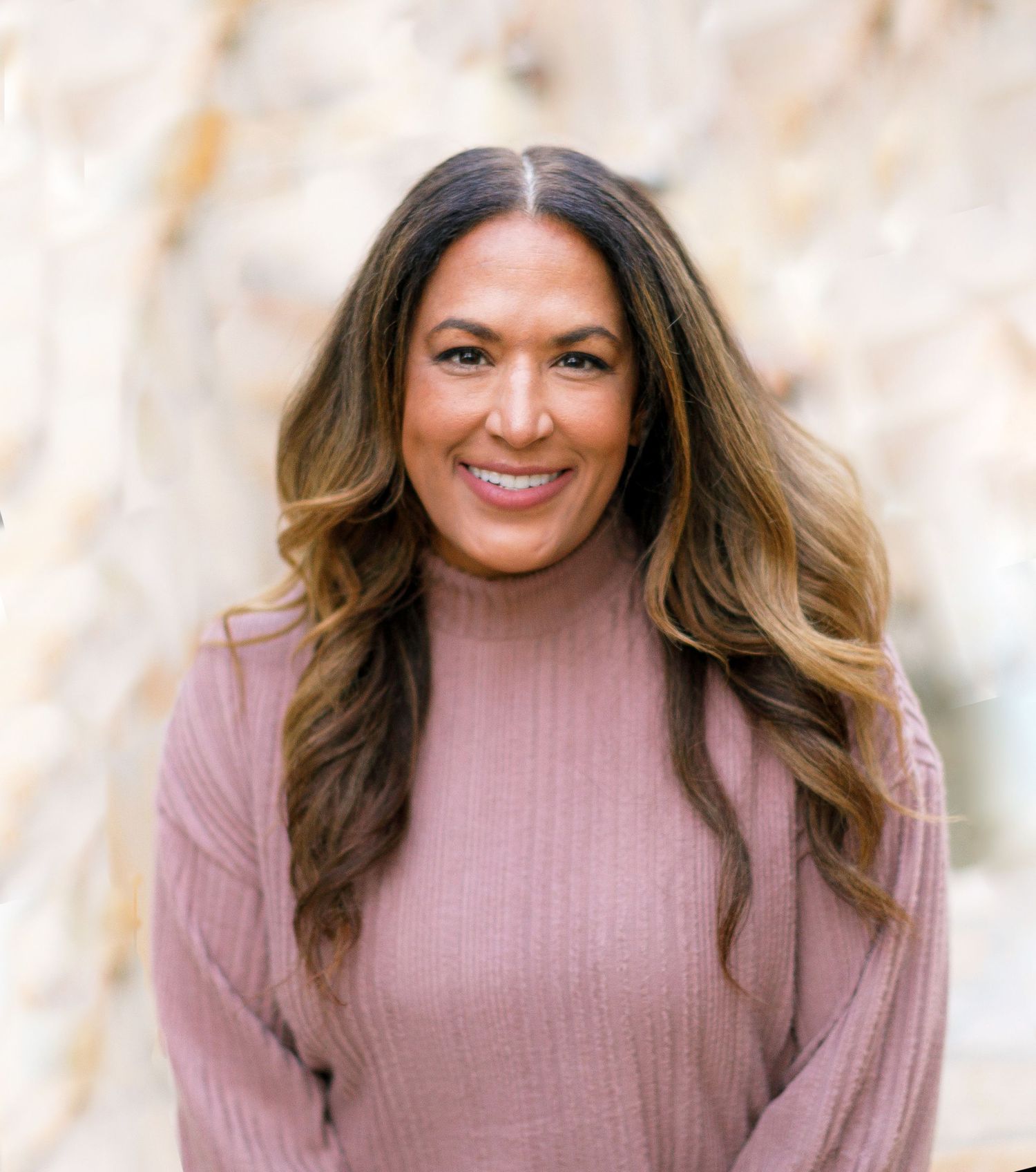 Smiling woman with long wavy brunette hair wearing a pink turtleneck sweater posing outdoors.