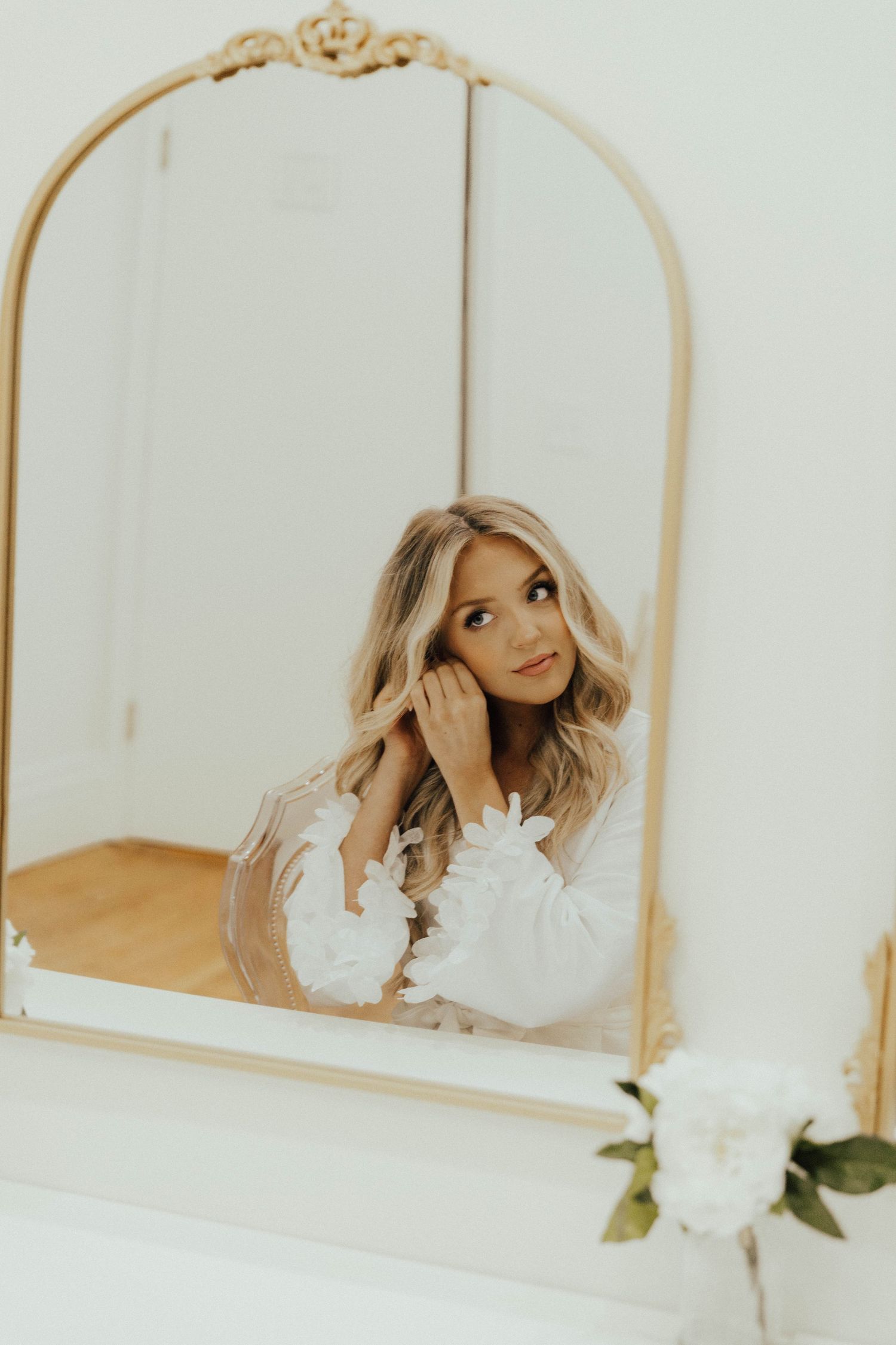 Reflection in ornate gold-framed mirror shows a figure in white dress with wavy blonde hair looking away dreamily.