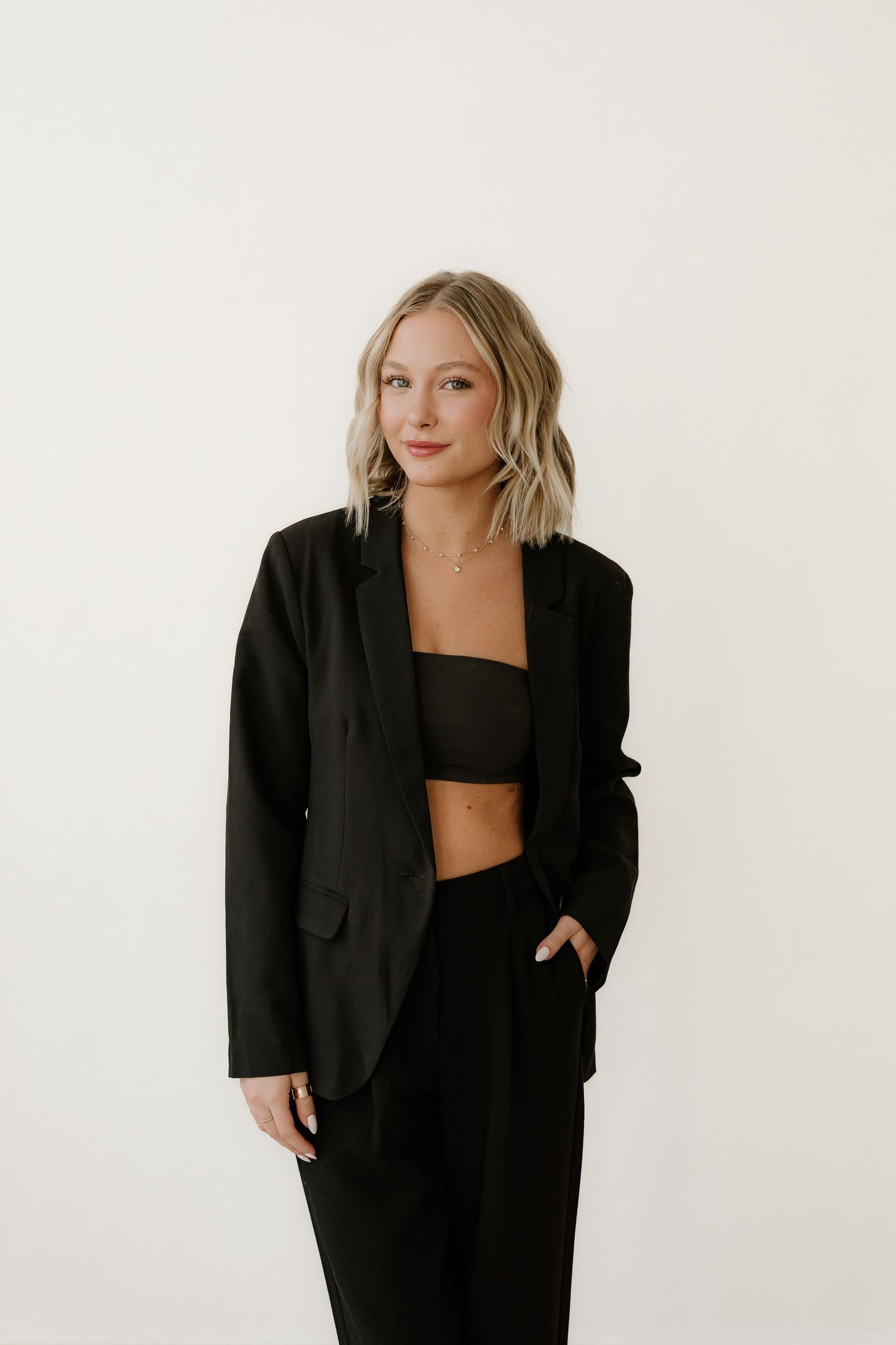 A person poses in a stylish black blazer, crop top and trousers against a white background.