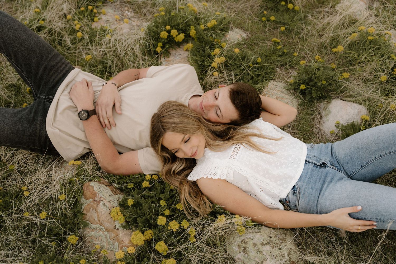 A couple lies together in a meadow with yellow wildflowers, wearing casual jeans and white tops.