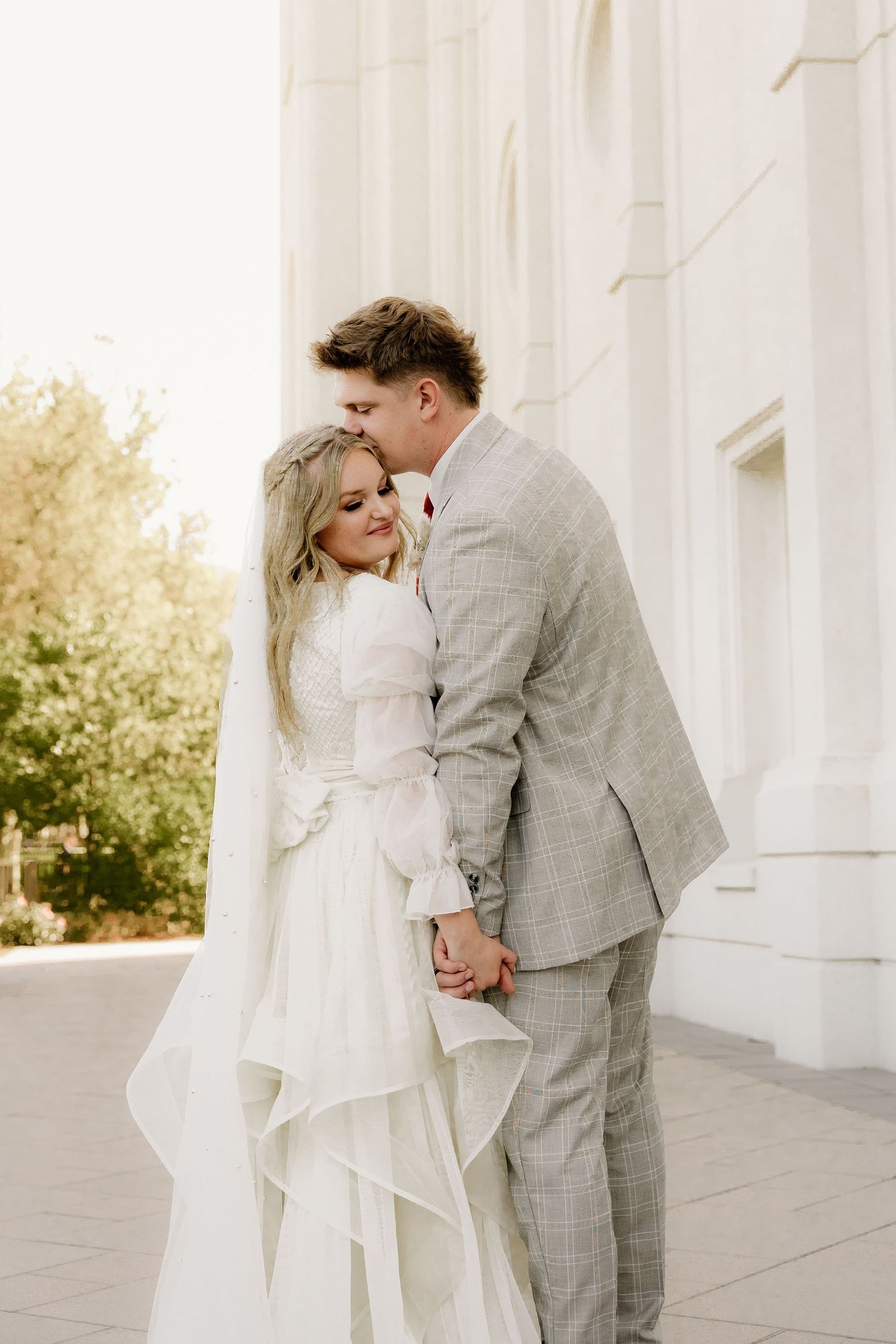 A romantic wedding couple in classic white dress and gray suit embrace outside a bright sunlit building.