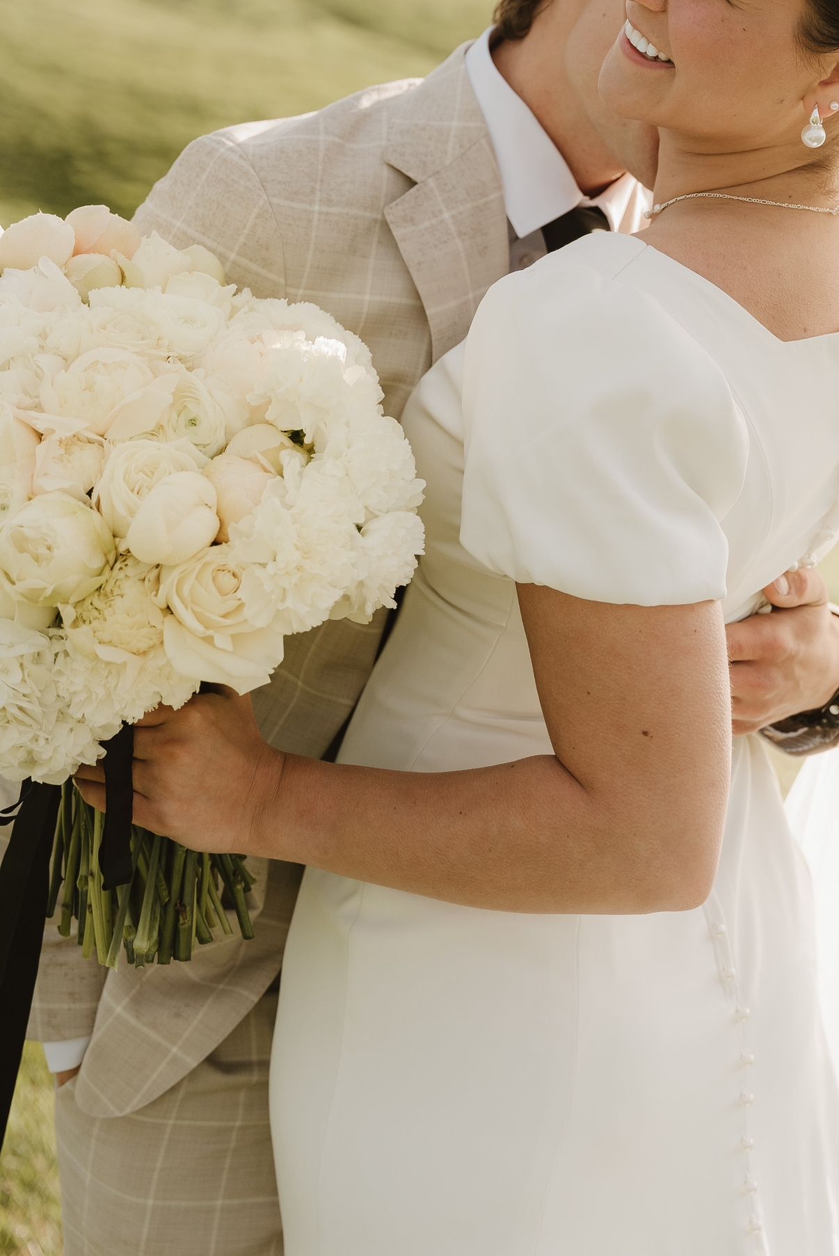 Close-up detail of bride in white dress holding elegant white rose bouquet next to groom in light checkered suit.