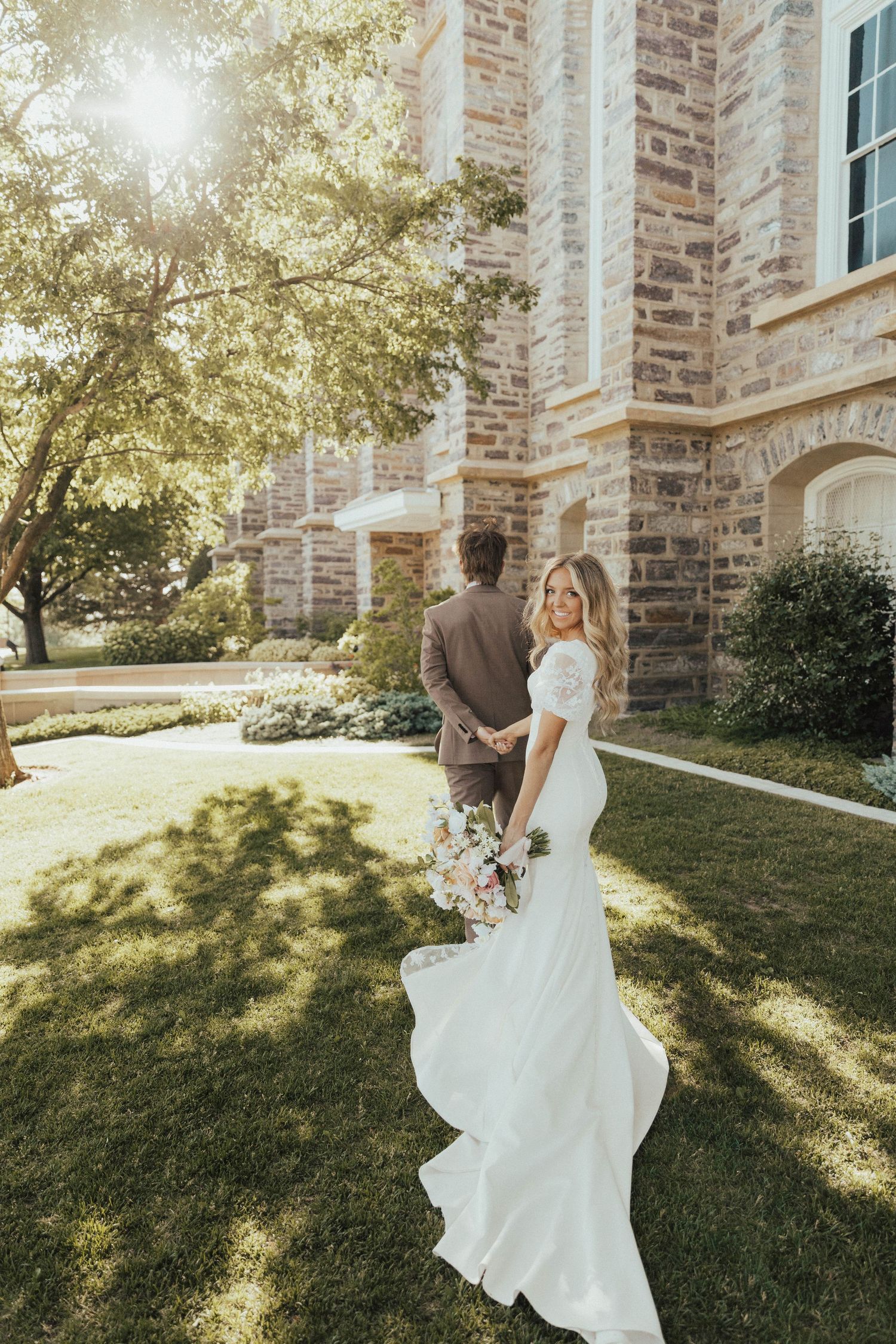 A bride in a flowing white dress holds a bouquet while standing outside a stone church building in golden sunlight.