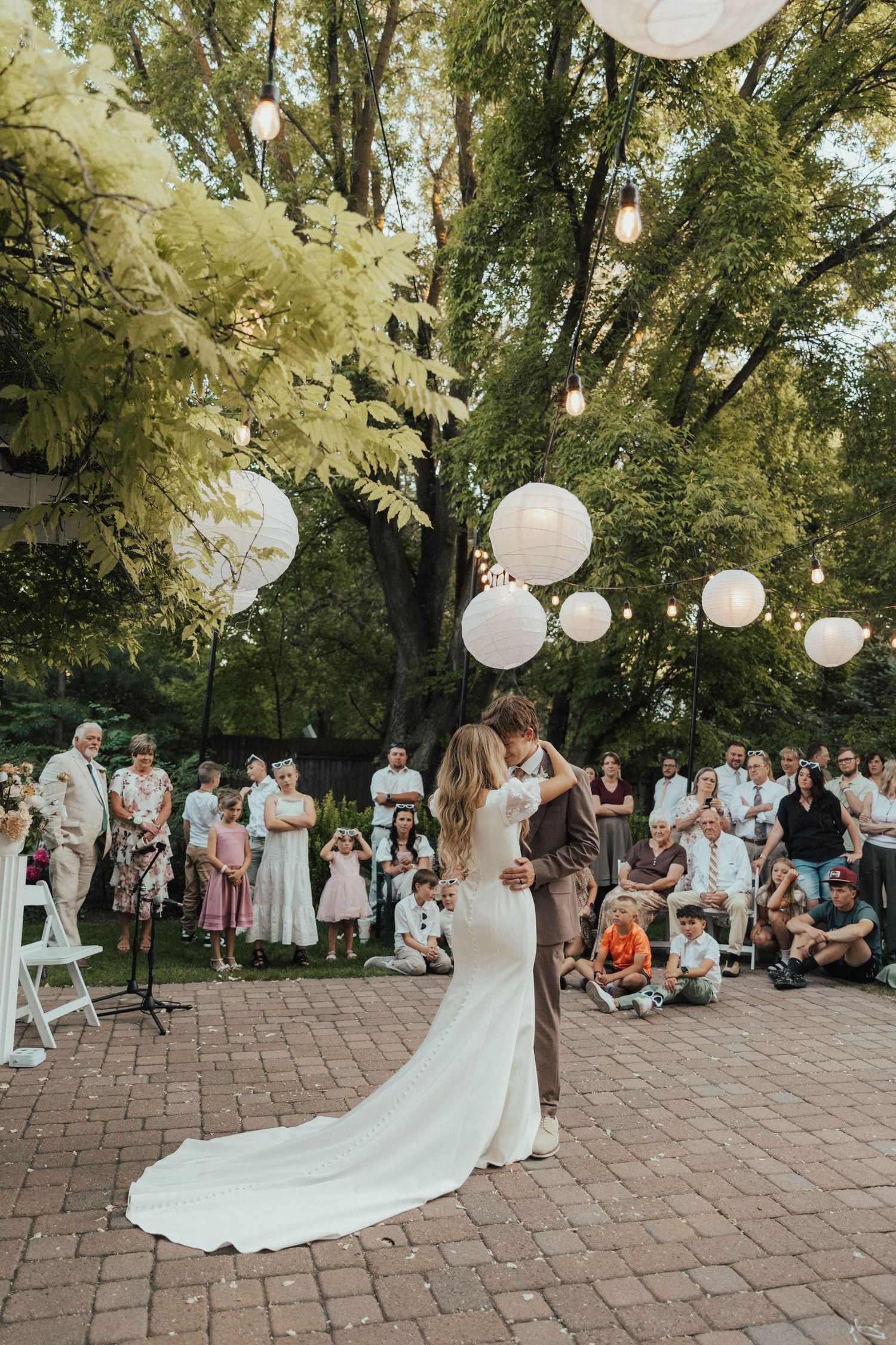 Romantic outdoor wedding reception with white paper lanterns hanging from trees above a brick patio surrounded by guests.