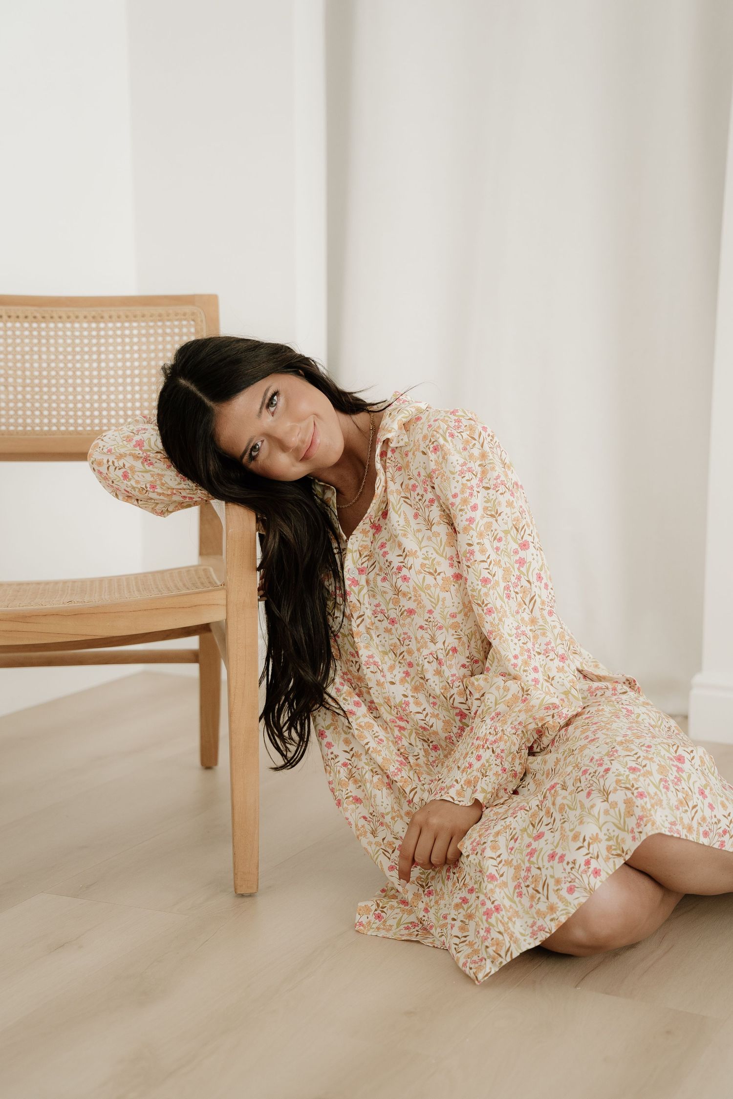 Woman in floral midi dress sitting on floor leaning against wooden rattan chair in bright minimalist room.