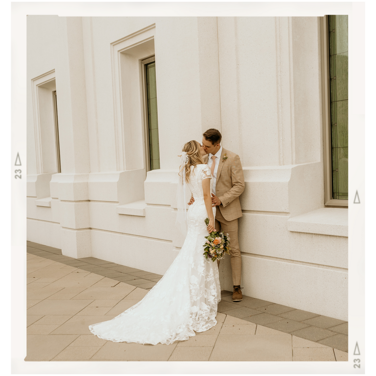 Wedding couple in elegant attire embrace against white columned building exterior.