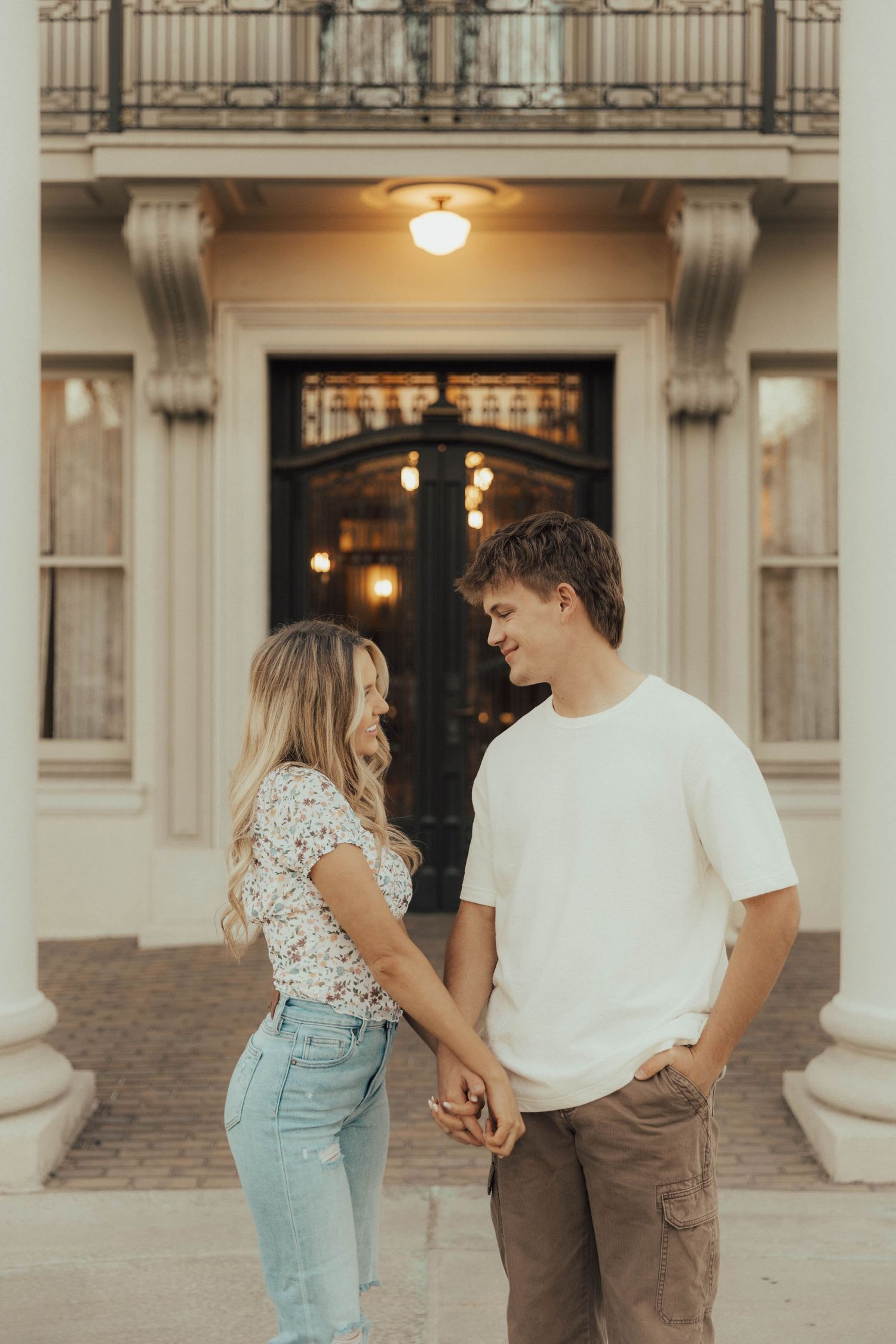 A couple dressed in casual clothing hold hands in front of an elegant building entrance at dusk.