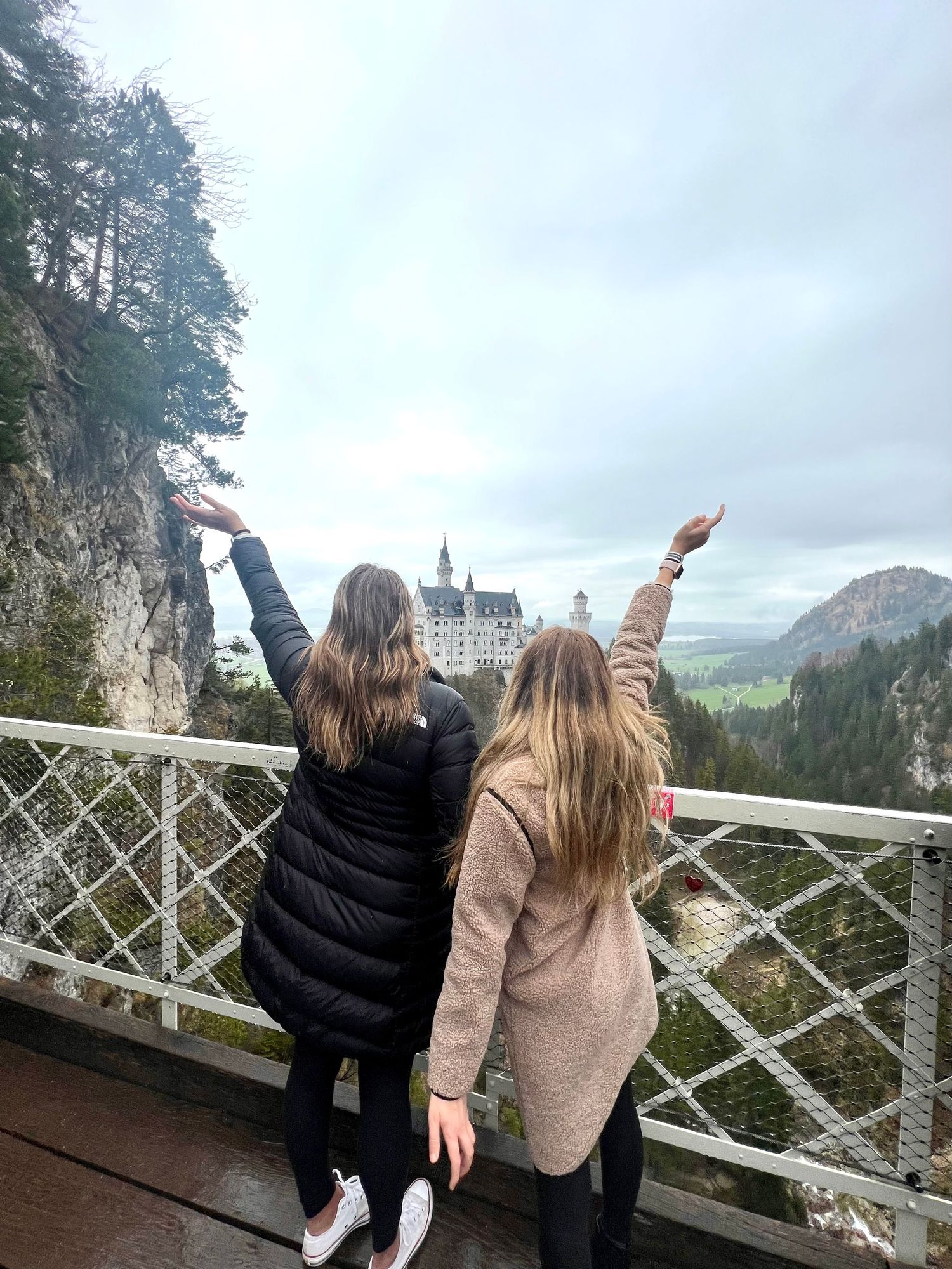 Two people strike poses on viewing platform overlooking Neuschwanstein Castle and Bavarian Alps on cloudy day.