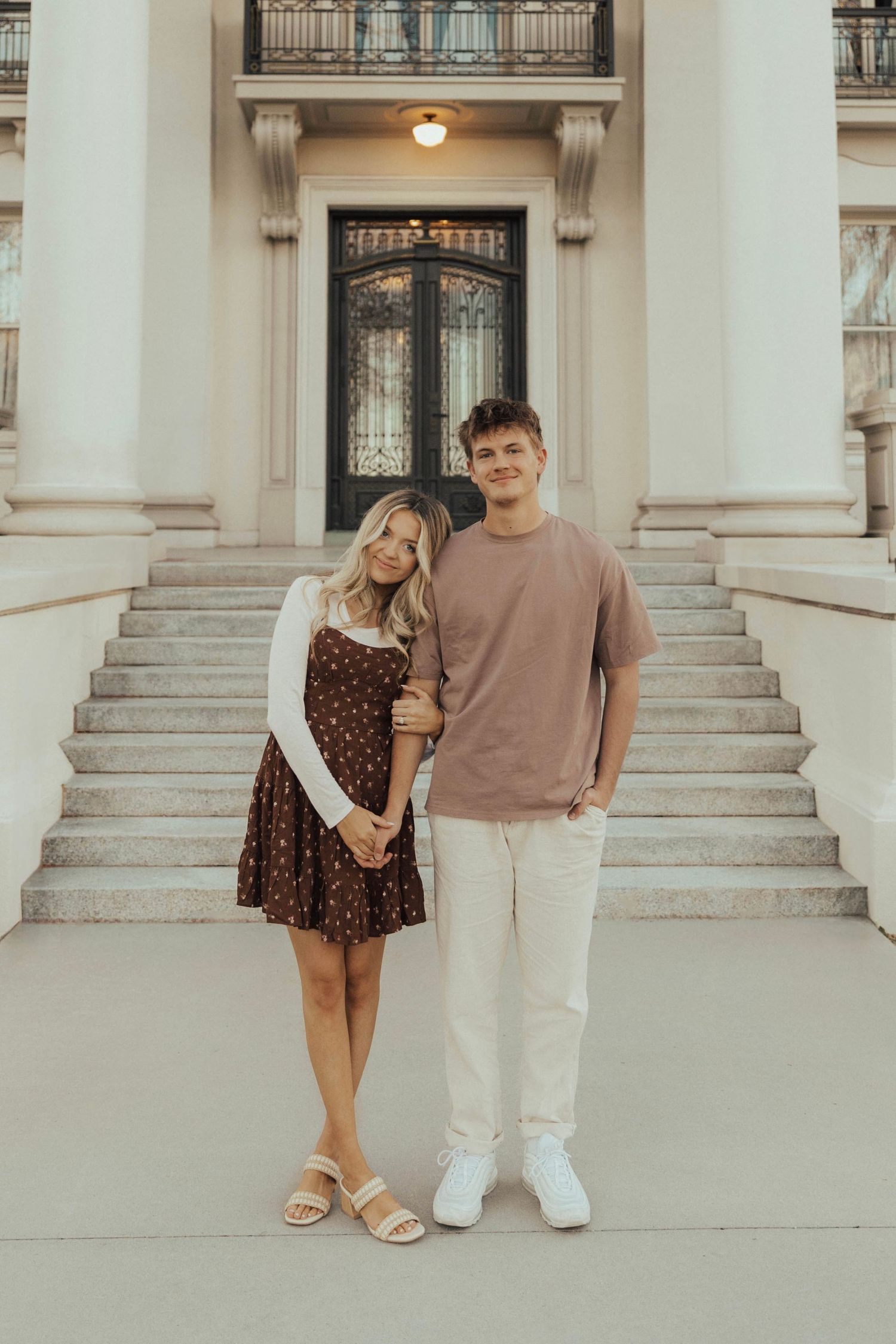 A couple stands on white marble steps in casual attire with an ornate doorway and columns behind them.