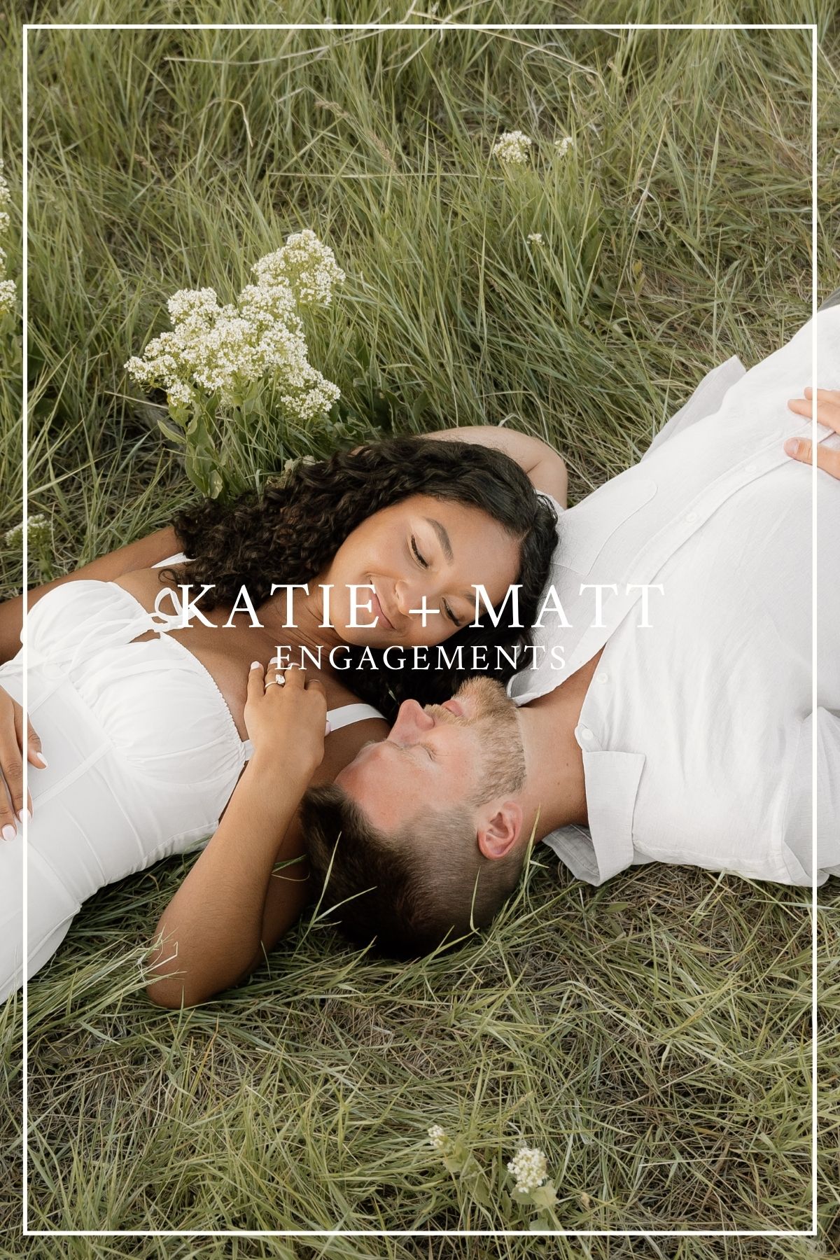 A couple lies in tall grass with white wildflowers during their romantic engagement photo session.